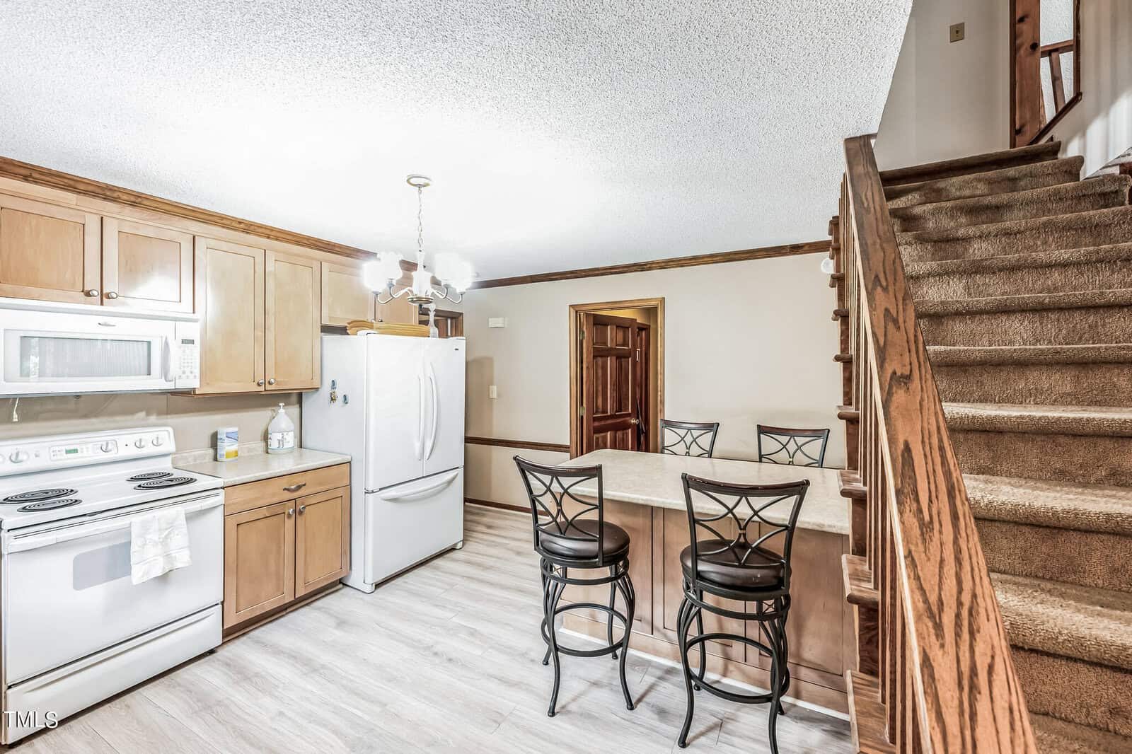 A modern kitchen with wooden cabinets, white appliances (stove, microwave, and refrigerator), a breakfast bar with three black barstools, light wood flooring, and a staircase leading upstairs. A wooden door and a chandelier are also visible.