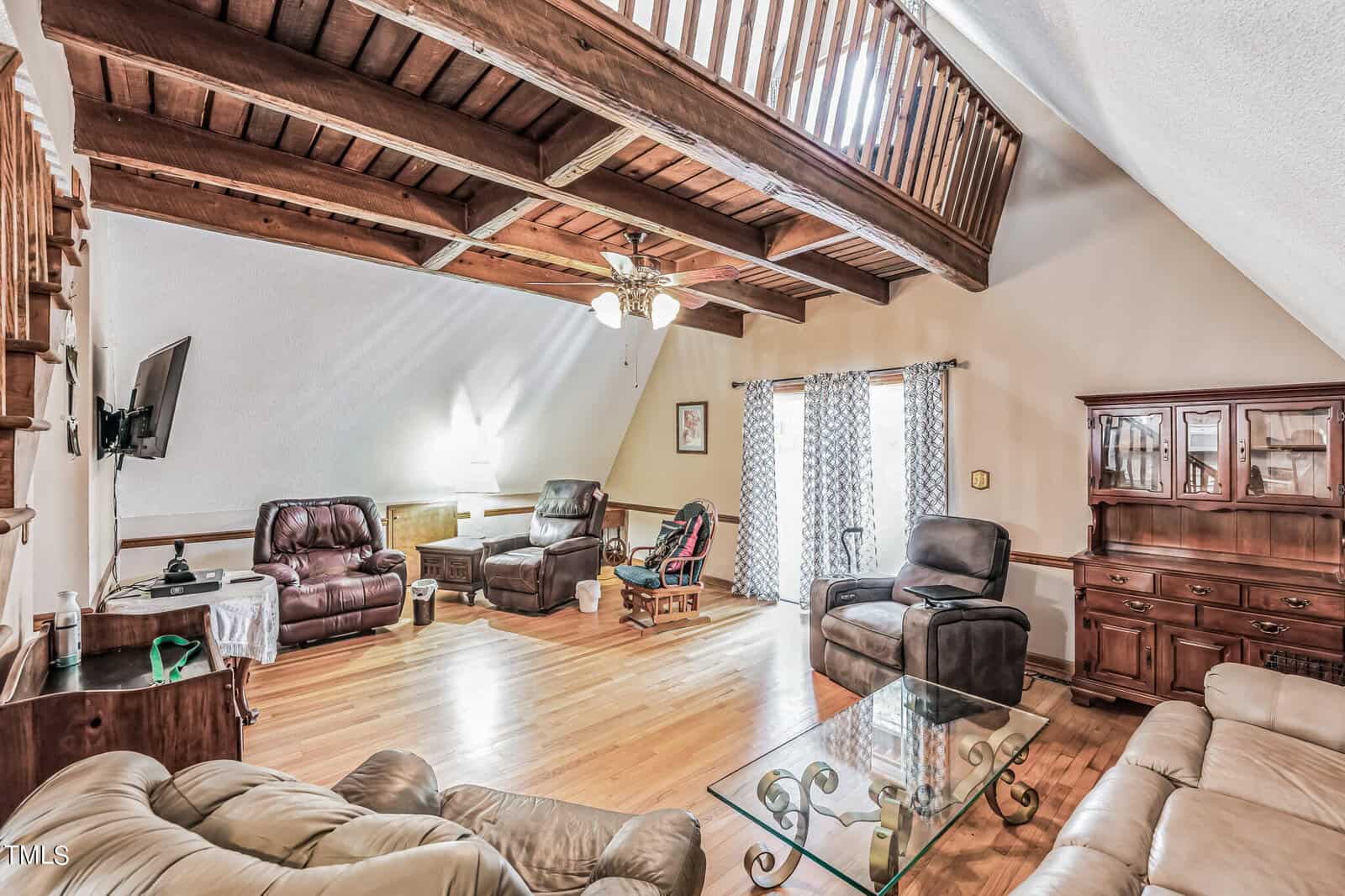 A spacious living room with wooden ceiling beams and a loft. The room features leather armchairs, a sofa, a glass coffee table, and a wooden cabinet. Large windows with patterned curtains allow natural light to brighten the space. A ceiling fan is mounted above.