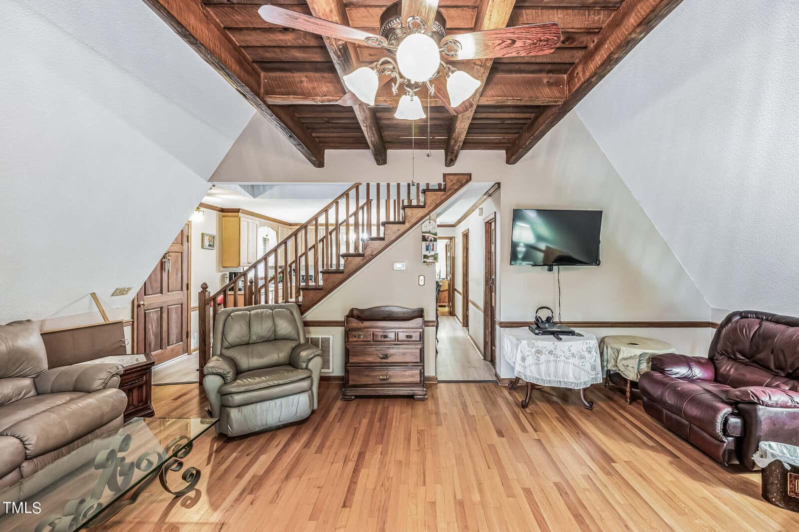 A cozy living room with wooden ceiling beams and a ceiling fan. The room features a flat-screen TV on the wall, an assortment of comfortable leather chairs and sofas, a wooden coffee table, and a staircase leading to the upper floor. The floor is light wood.