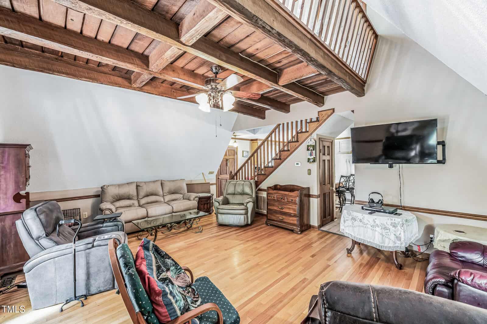 A spacious living room featuring wooden floors, exposed wooden ceiling beams, and a ceiling fan. There are various seating options including sofas and recliners. A TV is mounted on the wall above a table, and a wooden staircase with a railing leads to the upper floor.