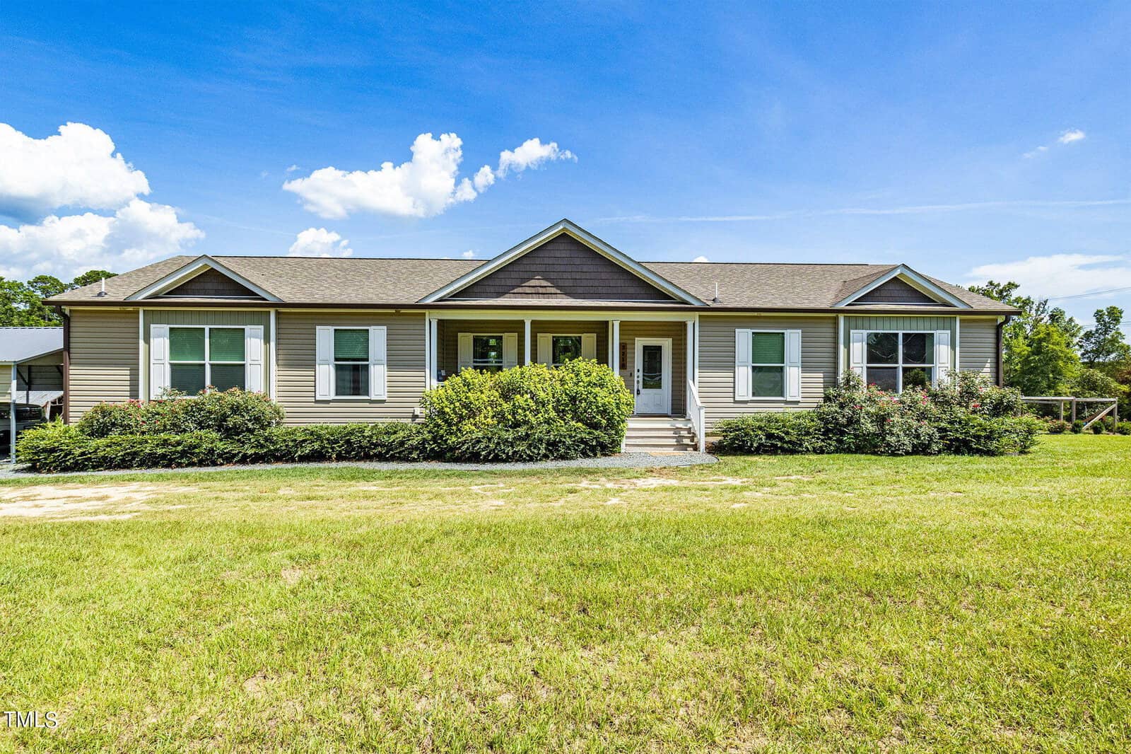A single-story house at 3218 Old Nc 75 features a gray exterior and green shutters, surrounded by neatly trimmed bushes and a grassy lawn. Under the clear blue sky with a few clouds, steps lead up to the front door, perfectly centered on the facade.