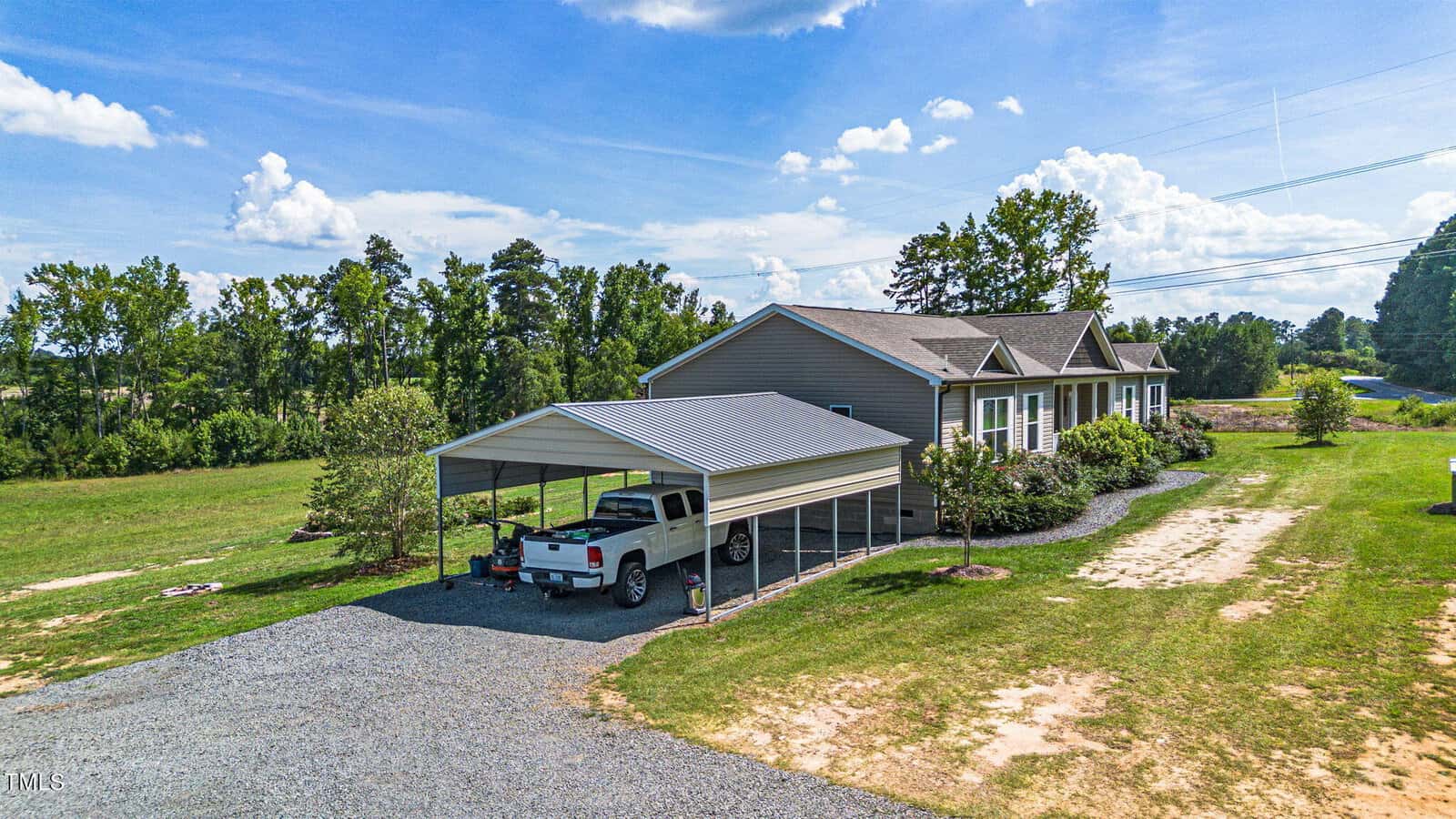 A single-story house with a light gray exterior is surrounded by green grass and trees. A white truck is parked under a metal carport beside the house on a gravel driveway. Clear blue sky and scattered clouds are overhead.