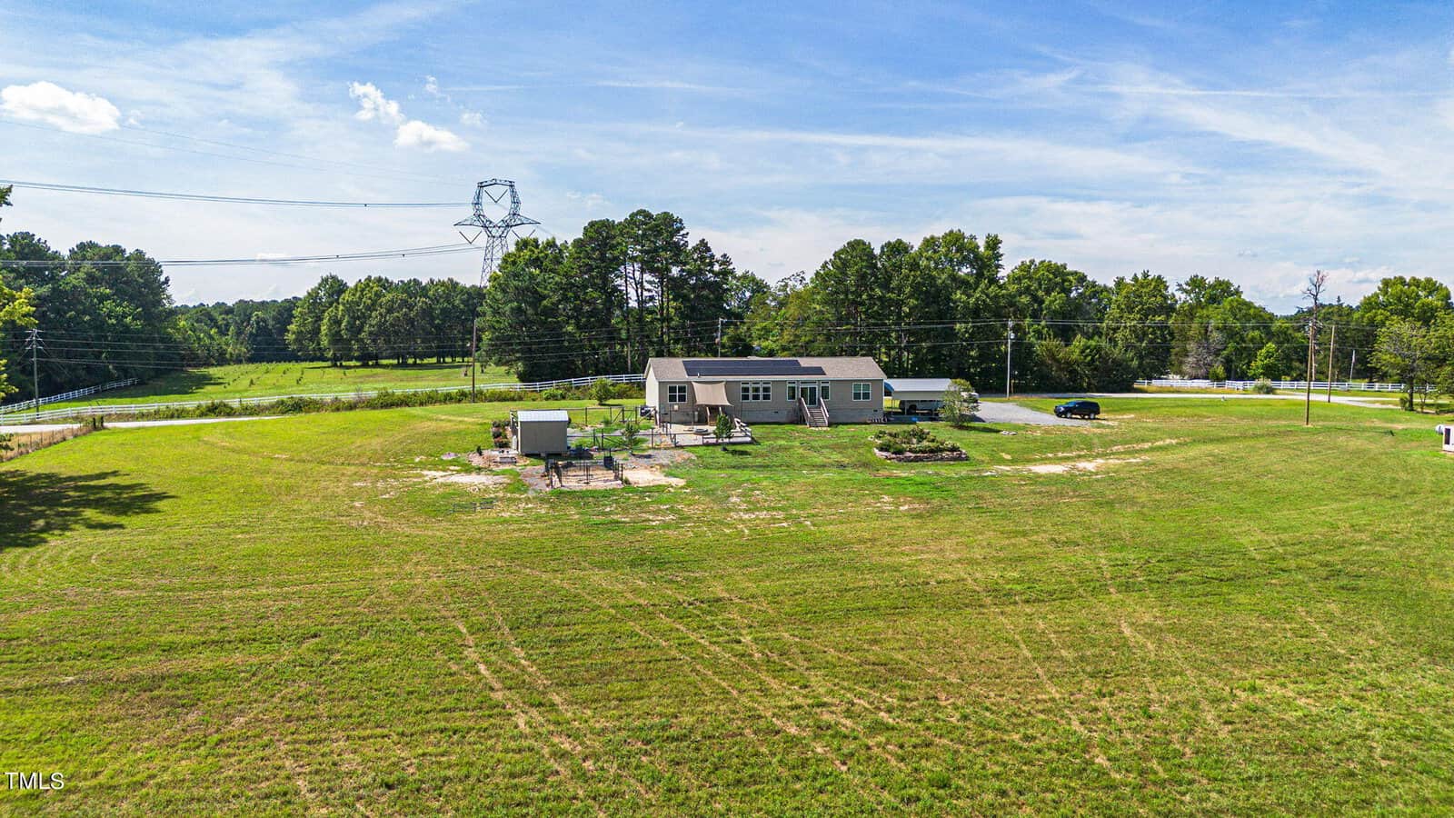 A spacious open field with a small house surrounded by trees. The house has a solar panel on the roof and several structures nearby. Power lines and a clear blue sky are visible in the background.