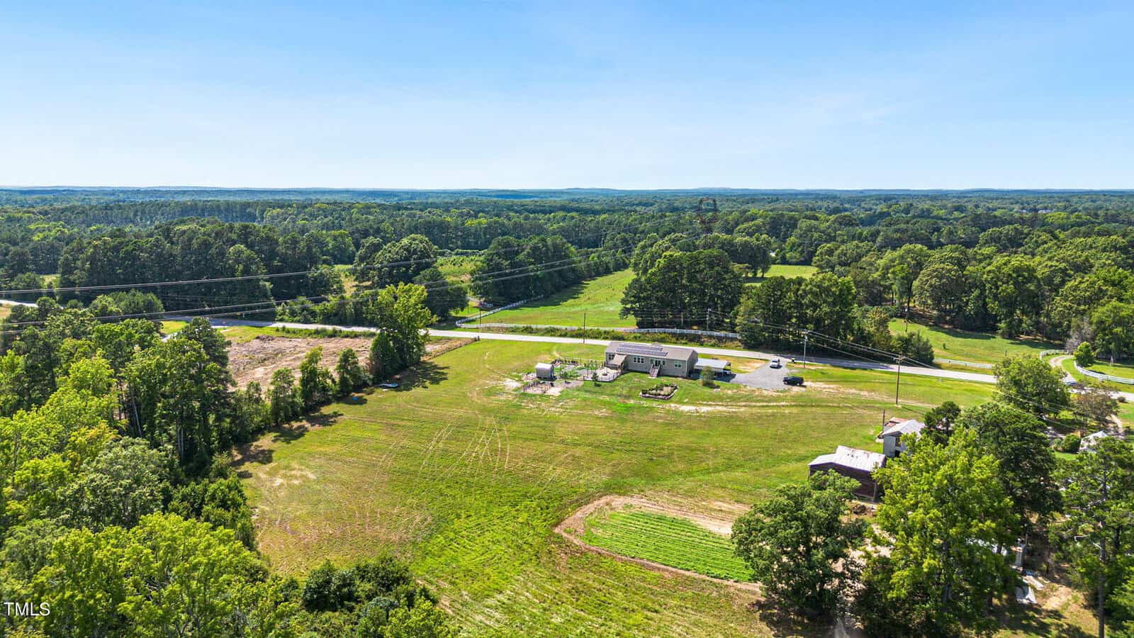 Aerial view of a countryside landscape featuring a long, single-story building surrounded by green fields and trees. A road runs nearby, and the horizon is visible under a clear blue sky.