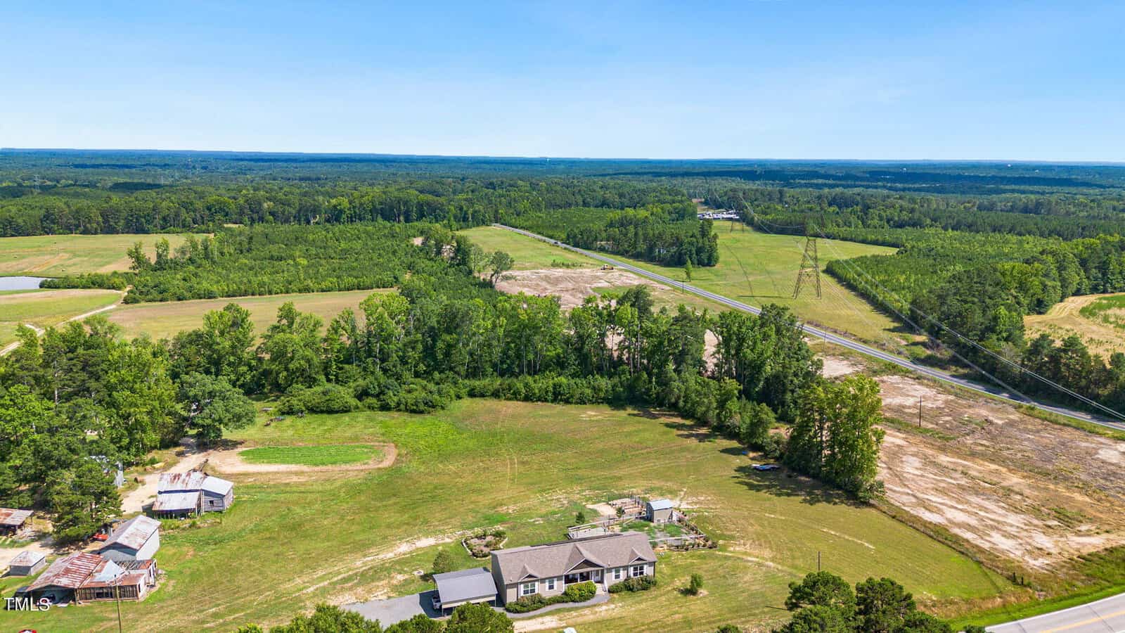 Aerial view of a rural landscape featuring a single house surrounded by fields and trees. A road cuts through the lush greenery, and the horizon showcases a vast expanse of forest under a clear blue sky.