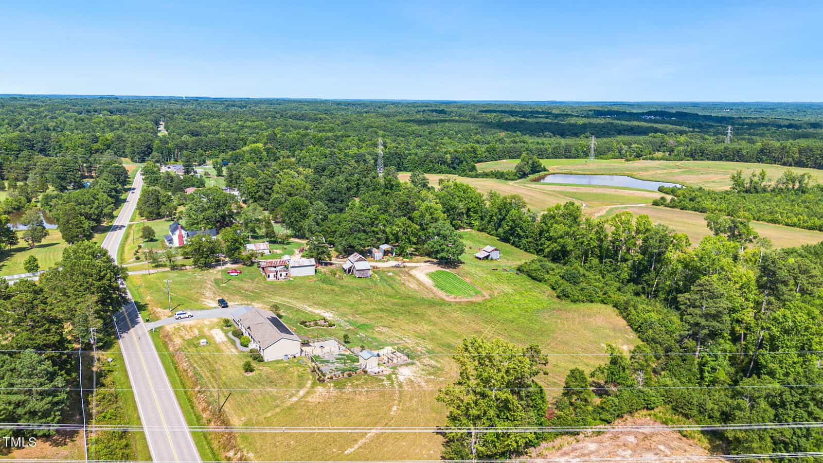 Aerial view of a rural landscape showing a cluster of houses and farm buildings surrounded by greenery and fields. A road runs parallel to the properties, with forested areas and a small pond visible in the distance under a clear blue sky.
