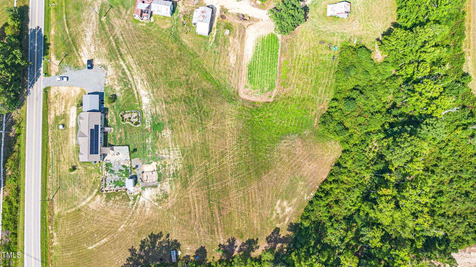 Aerial view of a rural landscape with a house and driveway at the left, surrounded by grassy fields and scattered trees. The right side of the image is densely forested, and several small structures are visible near the house.