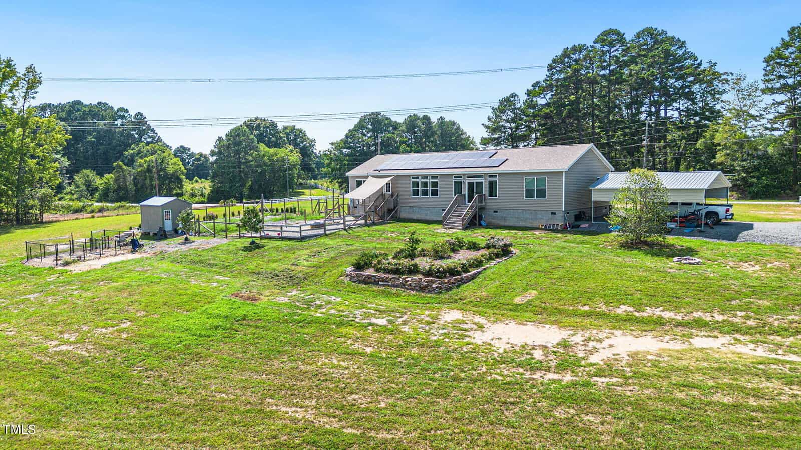 A modern single-story home with solar panels on the roof, surrounded by a large, grassy yard. A fenced garden area and a small shed are near the house. Tall trees and a blue sky are in the background.