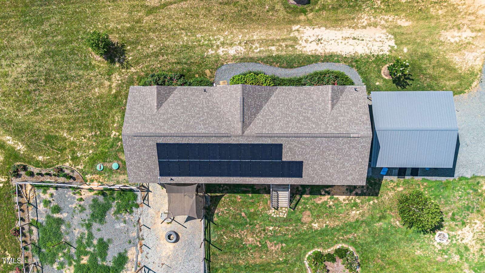 Aerial view of a house with solar panels on the roof. The yard features a defined pathway, a shaded seating area, and a small garden. The surrounding area is grassy with patches of dry ground.
