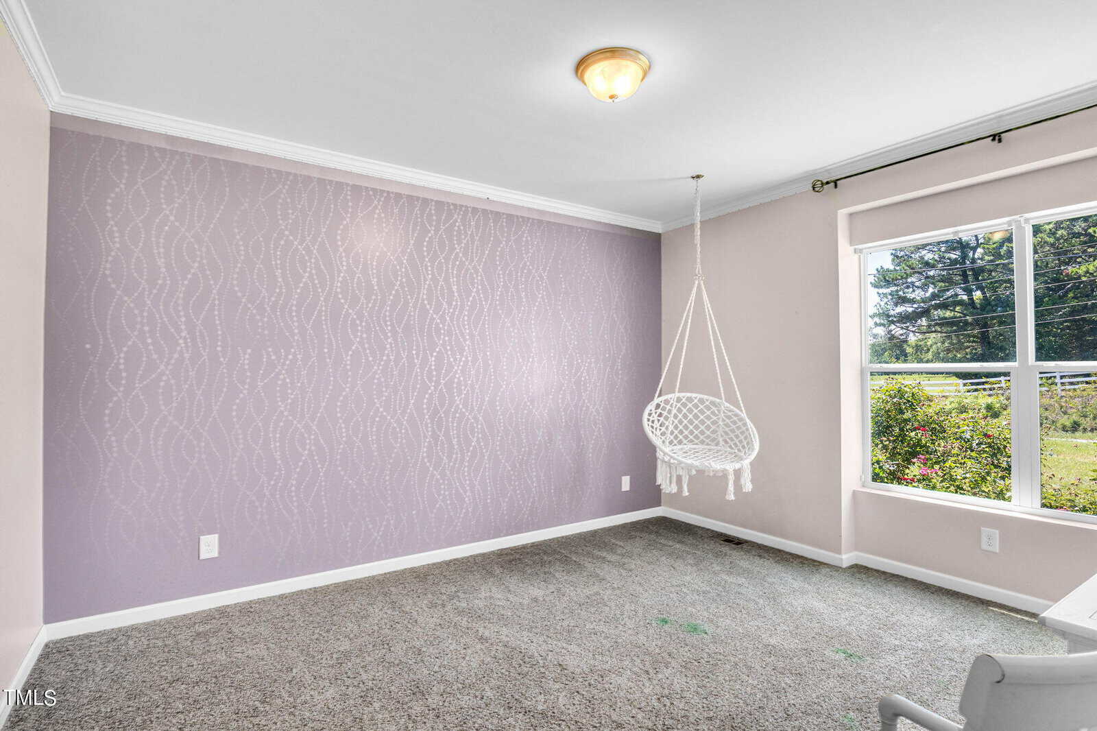A room with a purple accent wall featuring a subtle wavy pattern. A white hanging chair is suspended near a large window, which offers a view of greenery outside. The room has light gray carpet and a ceiling light fixture.