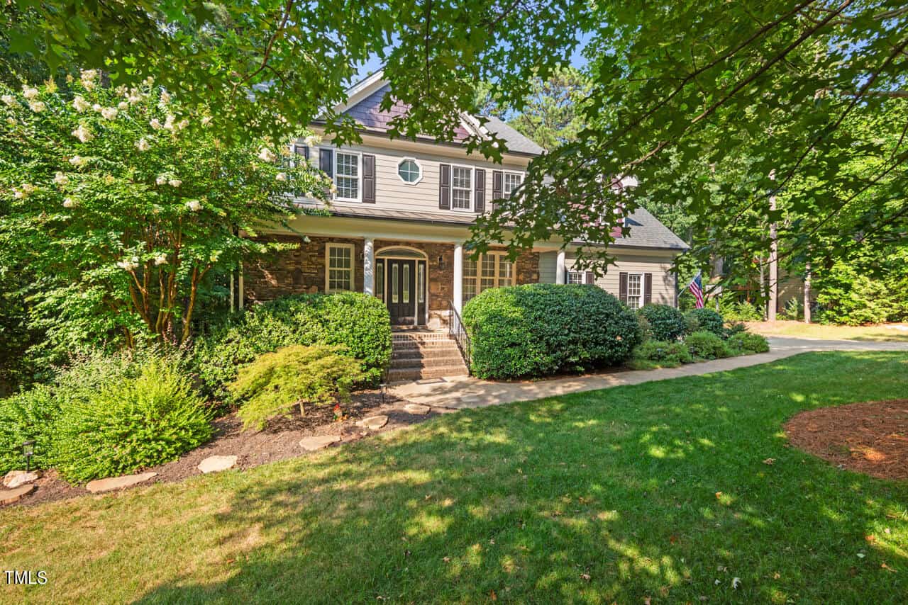 A two-story house with a dark front door, surrounded by lush greenery and trees. The exterior features a mix of stone and siding, and there is a well-maintained lawn with shrubs, a flag, and stepping stones leading to the entrance.