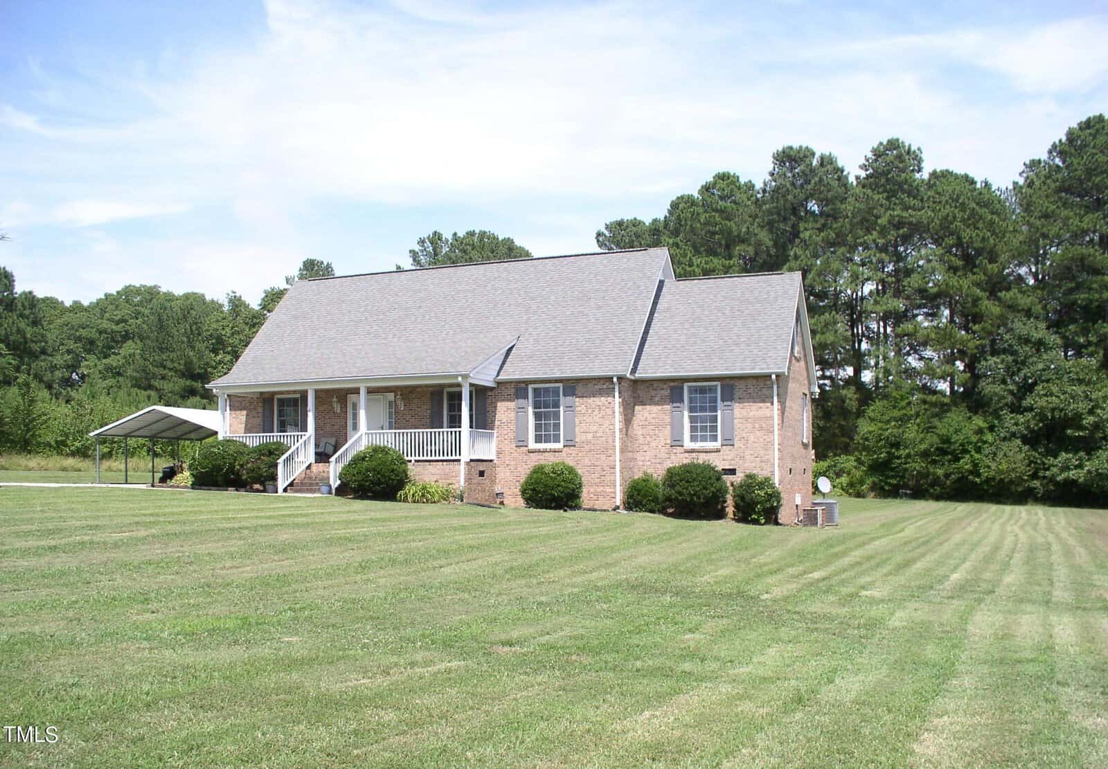 A brick house at 153 Cedar Grove Drive with a front porch, white railings, and a gray roof sits in the middle of a large, neatly mowed lawn in Henderson, NC. Bushes are planted along the house's foundation, and tall trees line the background. A small structure, possibly a carport, is on the left side of the house.