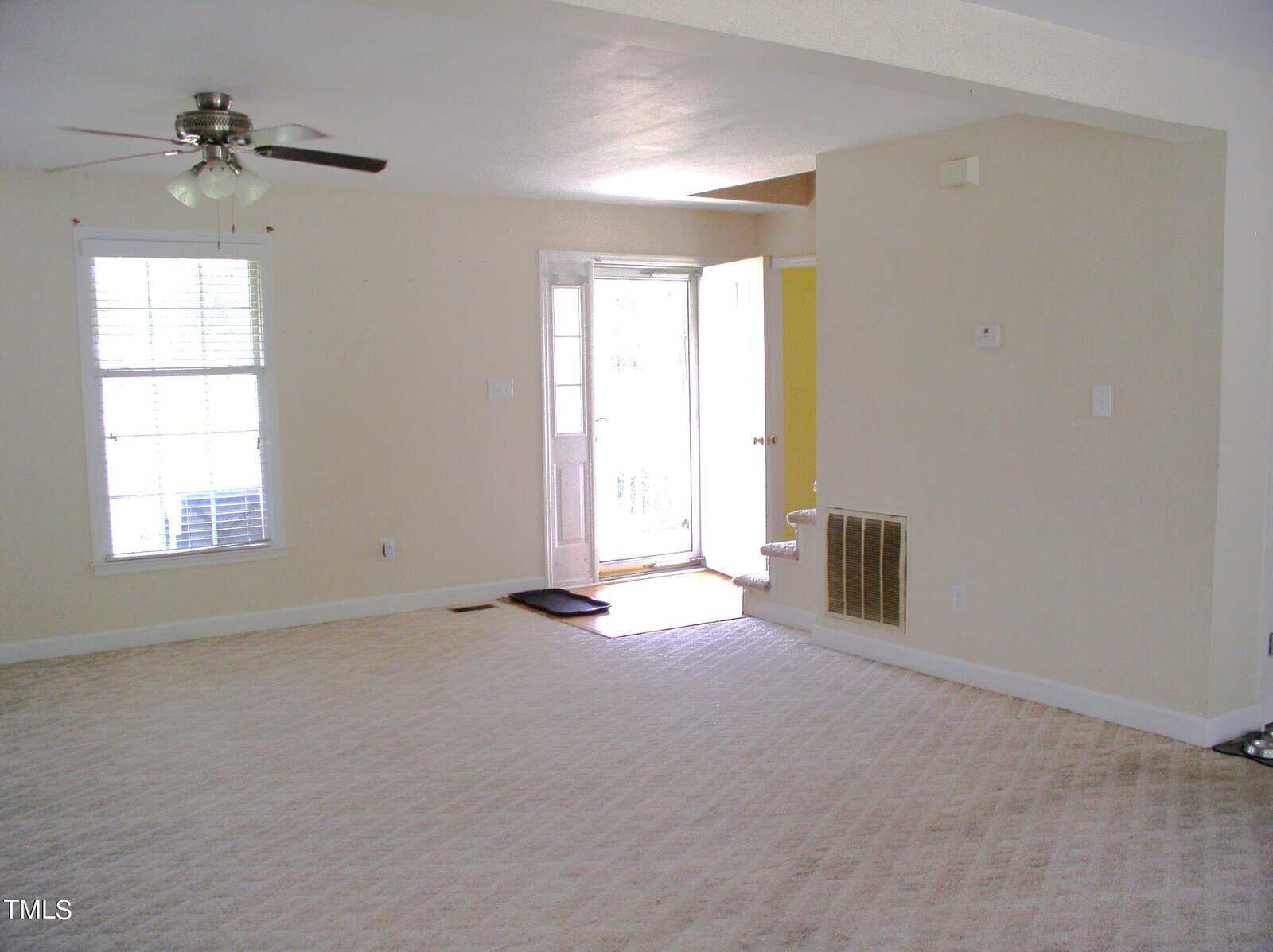 A spacious, empty living room with beige walls and carpet flooring. The room features a ceiling fan with light and a window with blinds on the left. An open doorway with steps and a mat leads to the outside. A vent is visible on one wall.