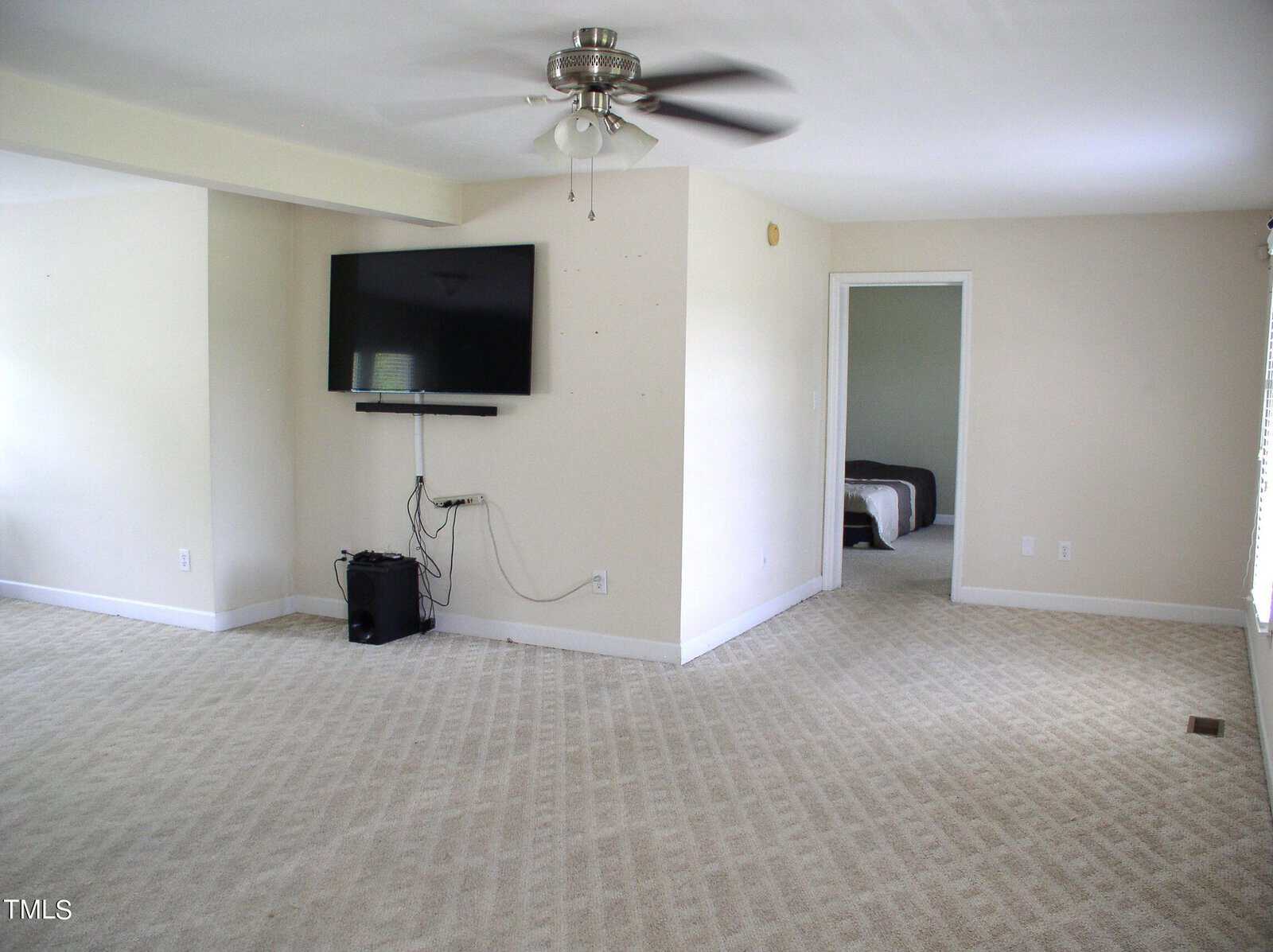 A spacious, carpeted room with a ceiling fan and a mounted flat-screen TV on the wall. Cables and a small black device rest on the floor below the TV. A doorway in the right wall leads to a bedroom with a visible bed. Light streams in from a window on the right.