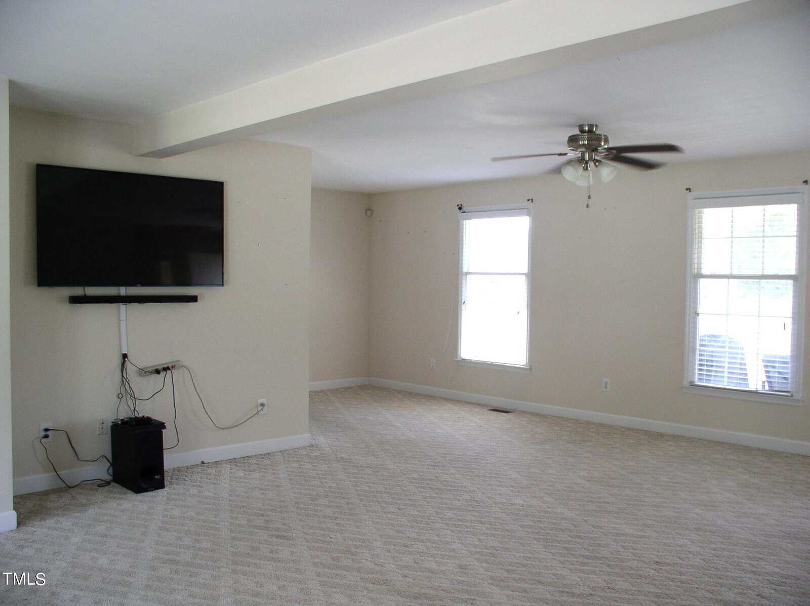 A sparsely furnished room with light beige walls and a carpeted floor. A large flat-screen TV is mounted on the wall with cables running down to a small electronic device on the floor. Two windows with blinds on the right and a ceiling fan above.