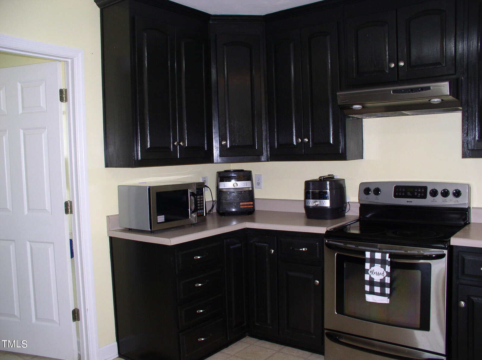 A kitchen with glossy black cabinets and beige countertops. On the counter are a microwave, an air fryer, and a pressure cooker. The kitchen features a stainless steel stove with a matching range hood. A white door and light yellow walls are visible.