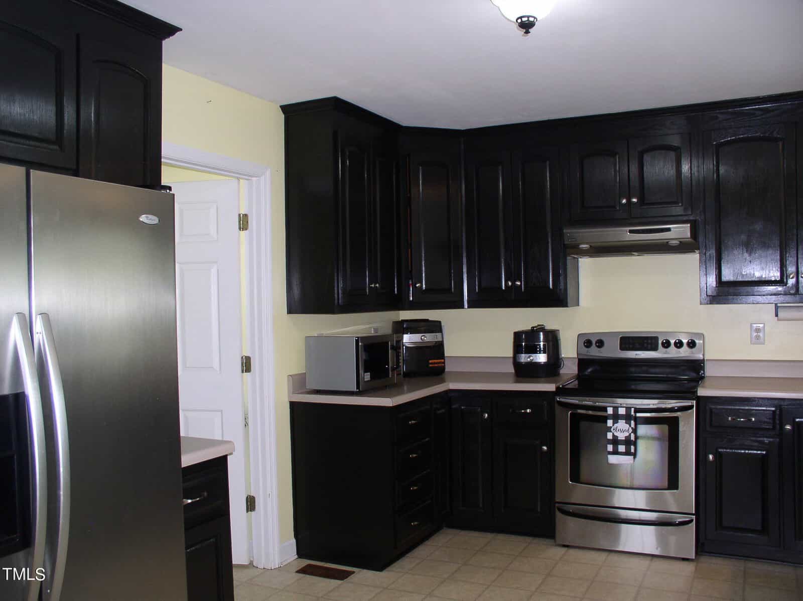 A modern kitchen features sleek black cabinets, stainless steel appliances including a refrigerator, stove, and oven, along with a microwave and air fryer on the countertop. Light yellow walls complement the dark cabinetry, and tiled flooring completes the look.