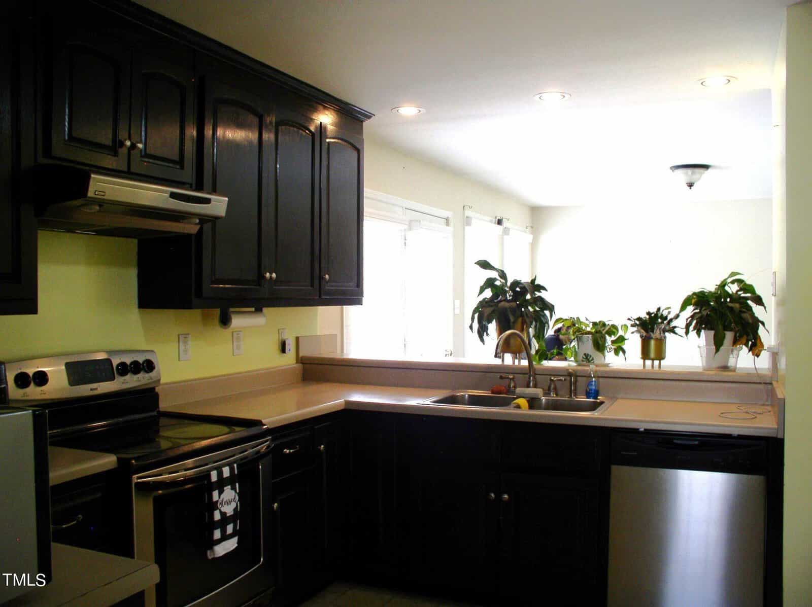 A modern kitchen featuring dark cabinetry, stainless steel appliances, and light countertops. There's a sink under a window with several potted plants on the windowsill. Recessed lighting illuminates the space, and a dishwasher is integrated into the cabinetry.