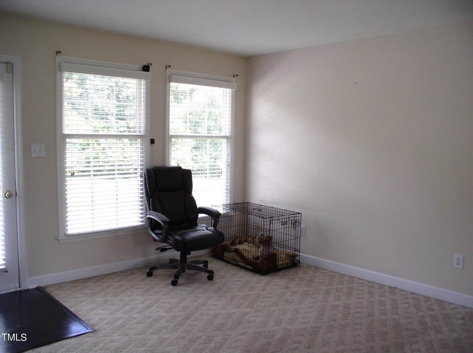 A minimally furnished room with beige carpet, off-white walls, and two large windows with blinds. The room contains a black office chair on wheels and a dog crate with a beige blanket inside. Light streams through the windows, providing natural illumination.