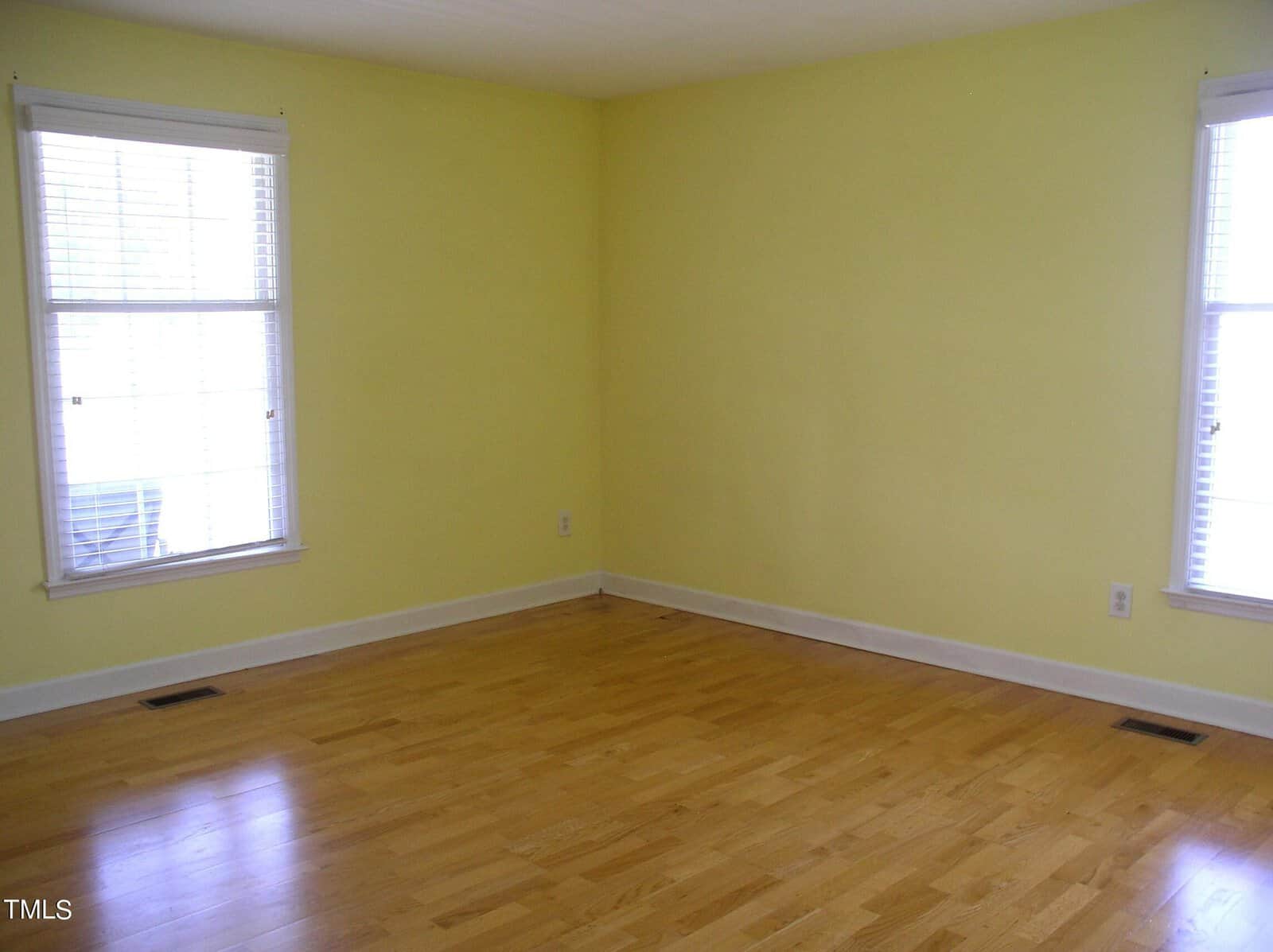 Empty room with light yellow walls, two large windows with white blinds, hardwood flooring, and white baseboards. The room is well-lit by natural light coming through the windows.