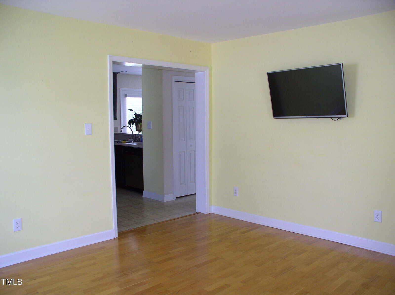 A mostly empty room with light yellow walls, light wood flooring, a wall-mounted TV, and a doorway leading to a kitchen with white cabinets and a window.