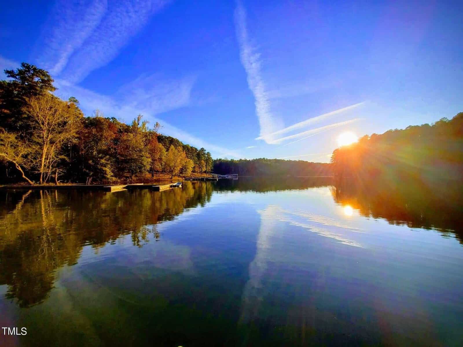 At sunrise on 3081 Jacksontown Road, a serene lake scene unfolds as trees line the shore. The sun casts a golden glow over the calm water, reflecting the vibrant blue sky and white contrails, creating a tranquil ambiance harmonized by Manson's clear, bright morning light.