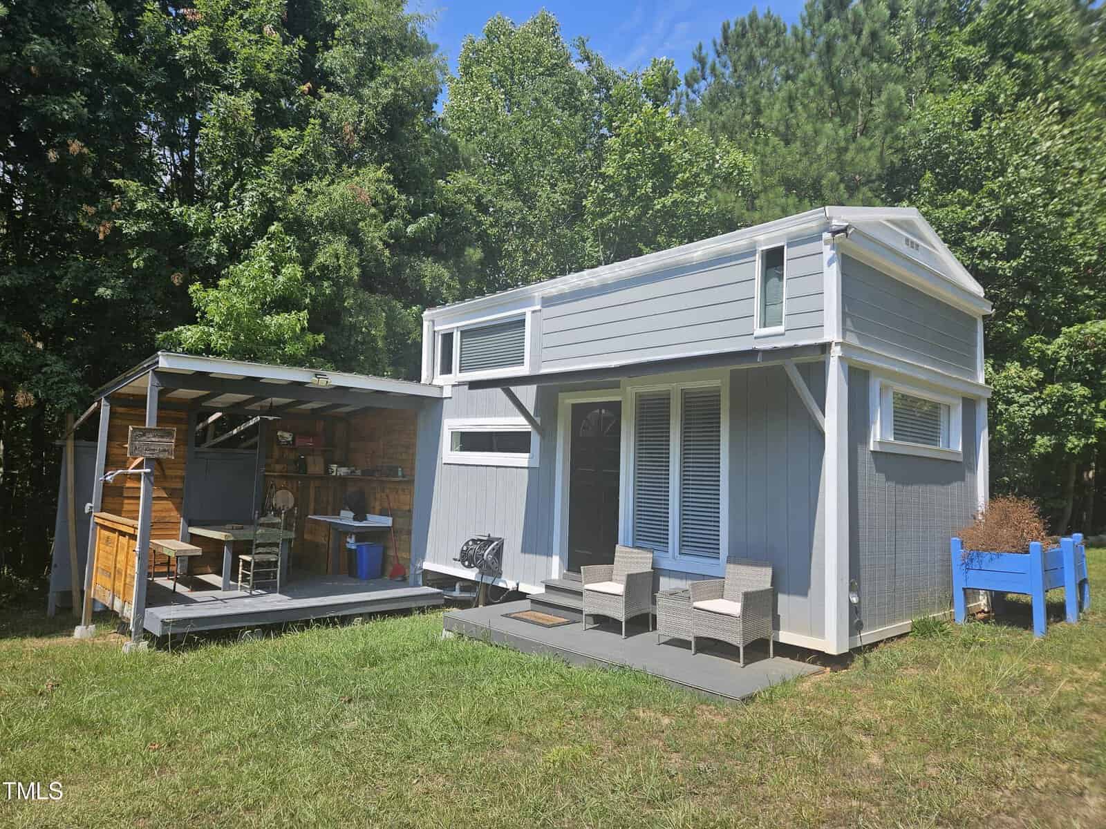 A modern, small gray and white tiny house with a covered outdoor kitchen area. The yard features a few chairs on the deck and a blue planter box. Trees surround the structure, providing a secluded setting.