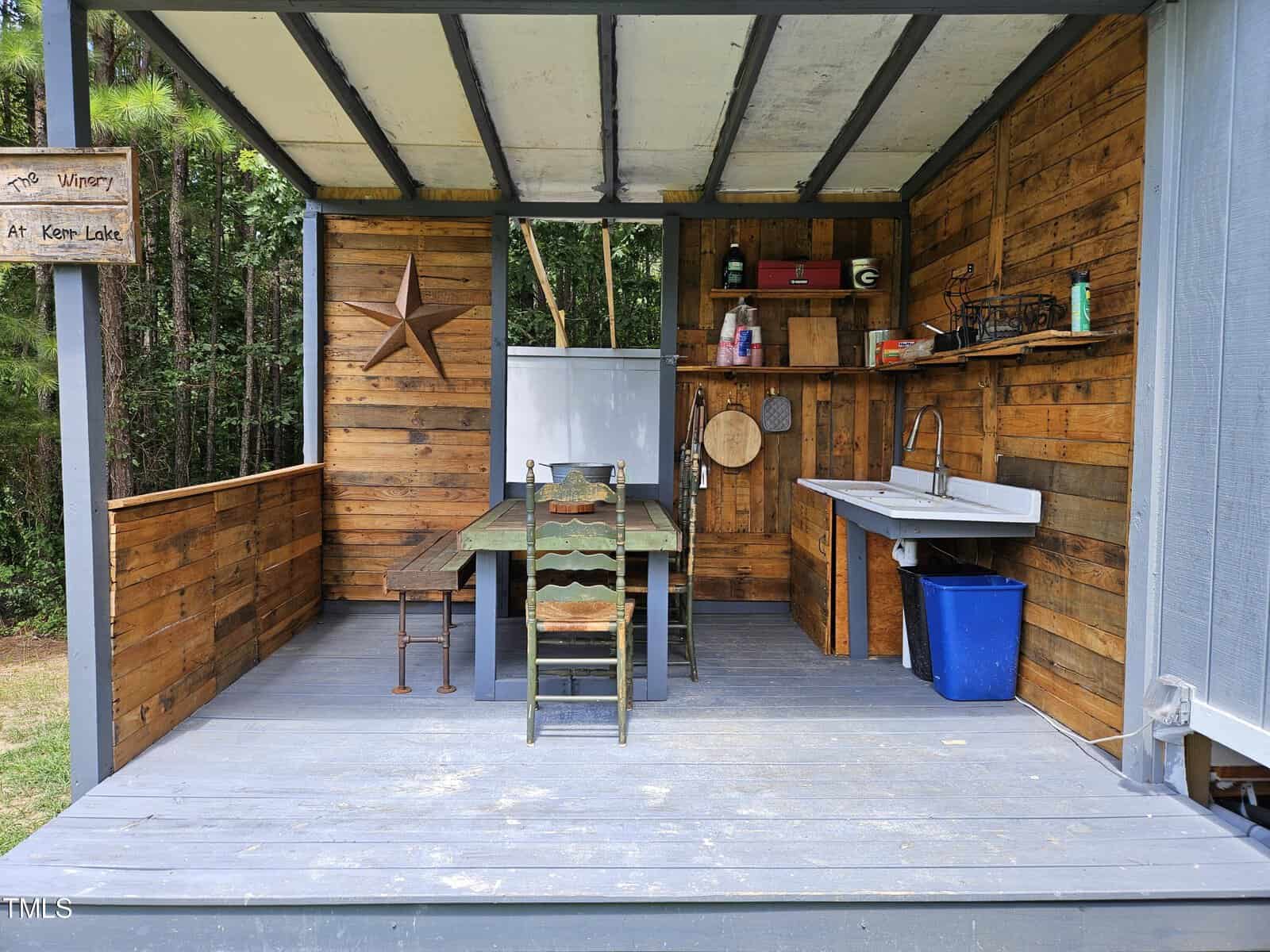 An outdoor kitchen with rustic wooden walls, a dining table with chairs, a large star decoration, and a sink. Shelves hold various items, and a blue trash bin is nearby. The setting is surrounded by greenery.