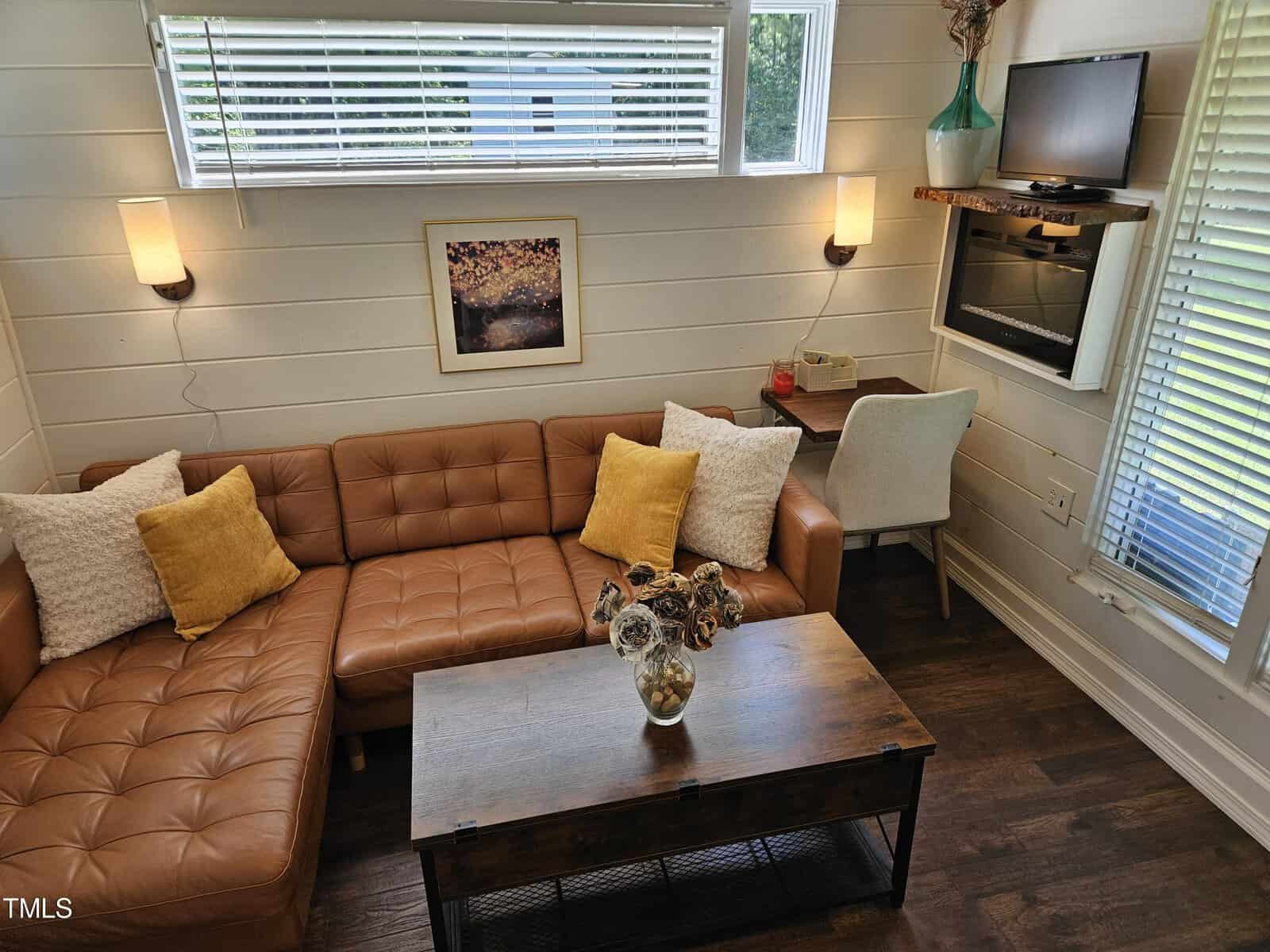 Cozy living room with a brown leather sectional sofa, assorted pillows, and a wooden coffee table. A small desk with a chair is against the wall. There are two wall sconces, a painting, and a small TV mounted above the desk. Natural light filters in through the blinds.