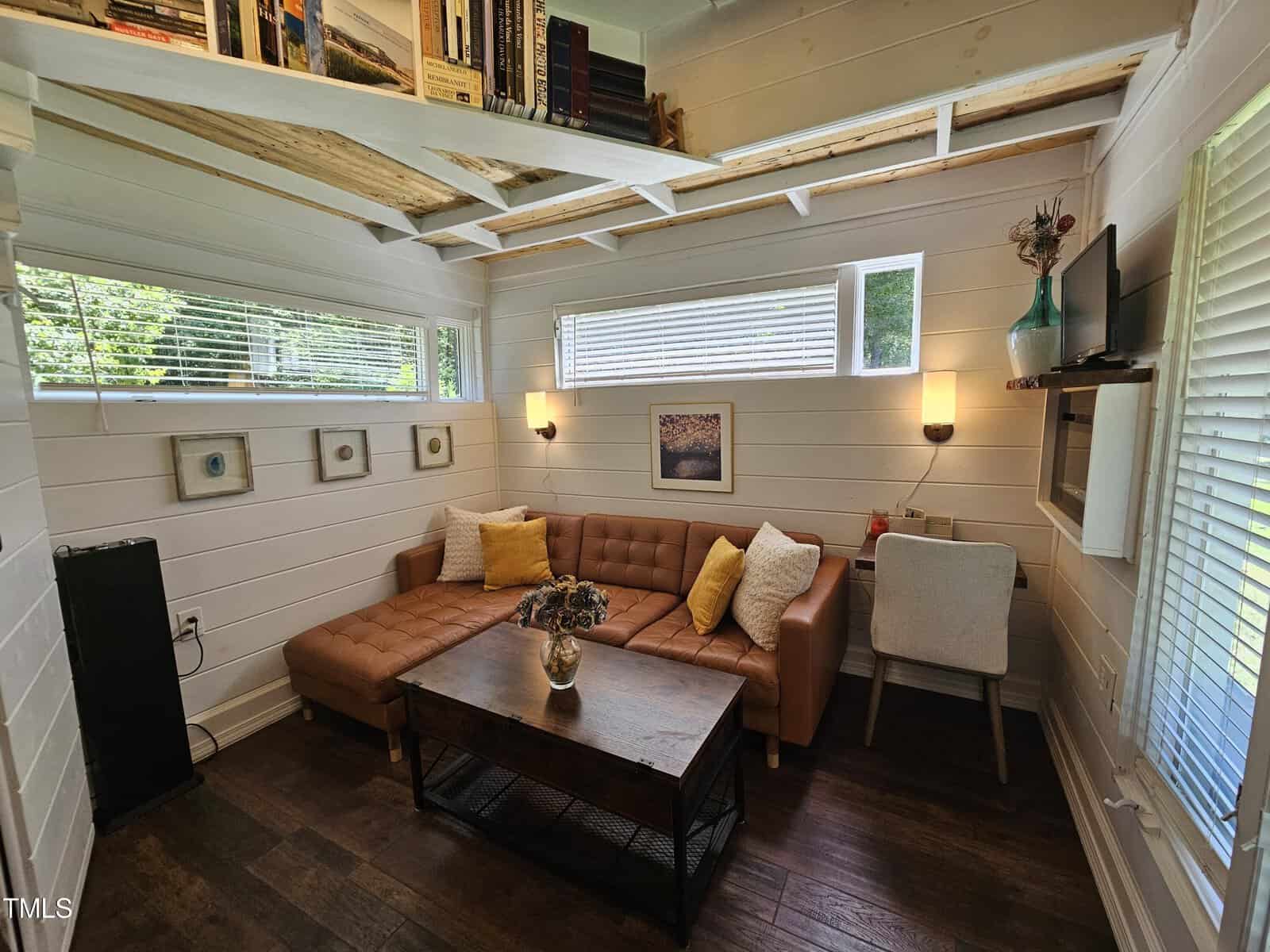 A cozy living room with white shiplap walls, a brown sectional sofa, a wooden coffee table, and wall-mounted shelves holding books. Two windows bring in natural light, and a small work desk is placed against one wall.