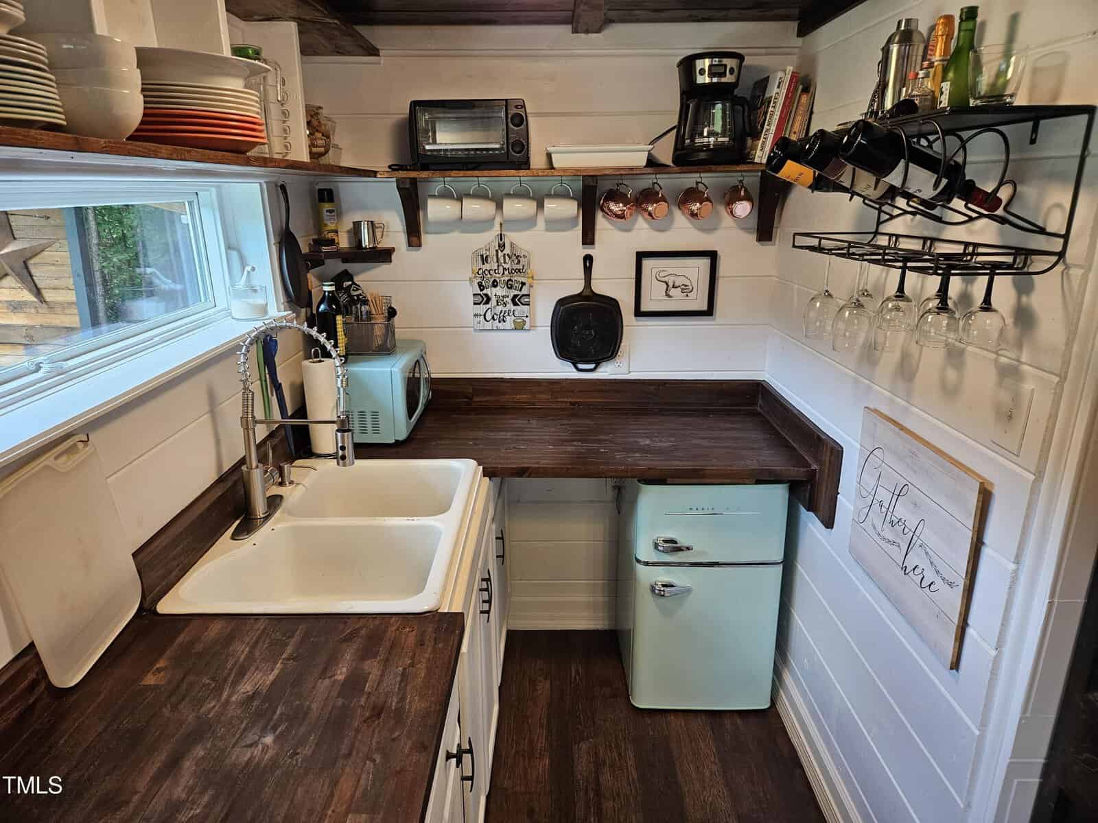 A cozy kitchen with wooden countertops and white cabinets. It features a small fridge, sink, microwave, coffee maker, and wall-mounted wine and dish racks. A window above the sink provides natural light to the tidy space.