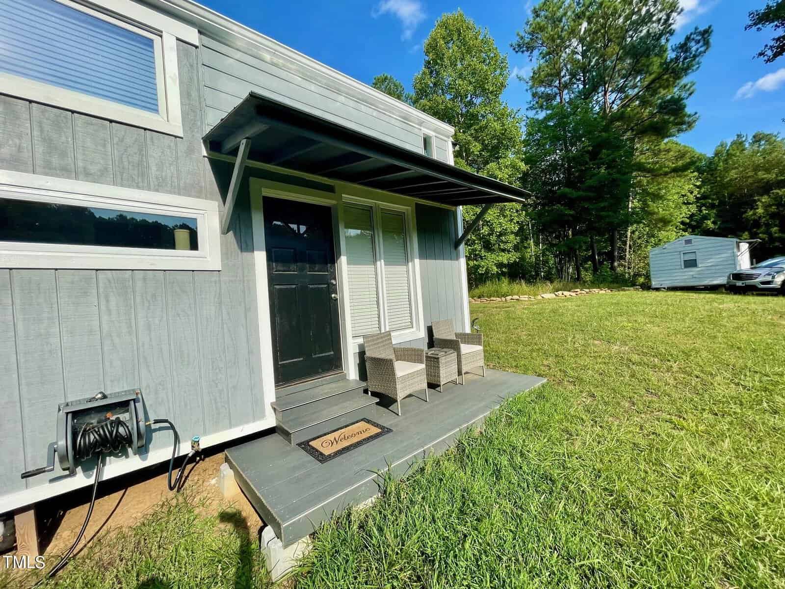 Small gray house with a black door and two wicker chairs on the porch. A "Welcome" mat is in front of the door. Surrounded by grass and trees, there's also a white trailer in the background under a blue sky.
