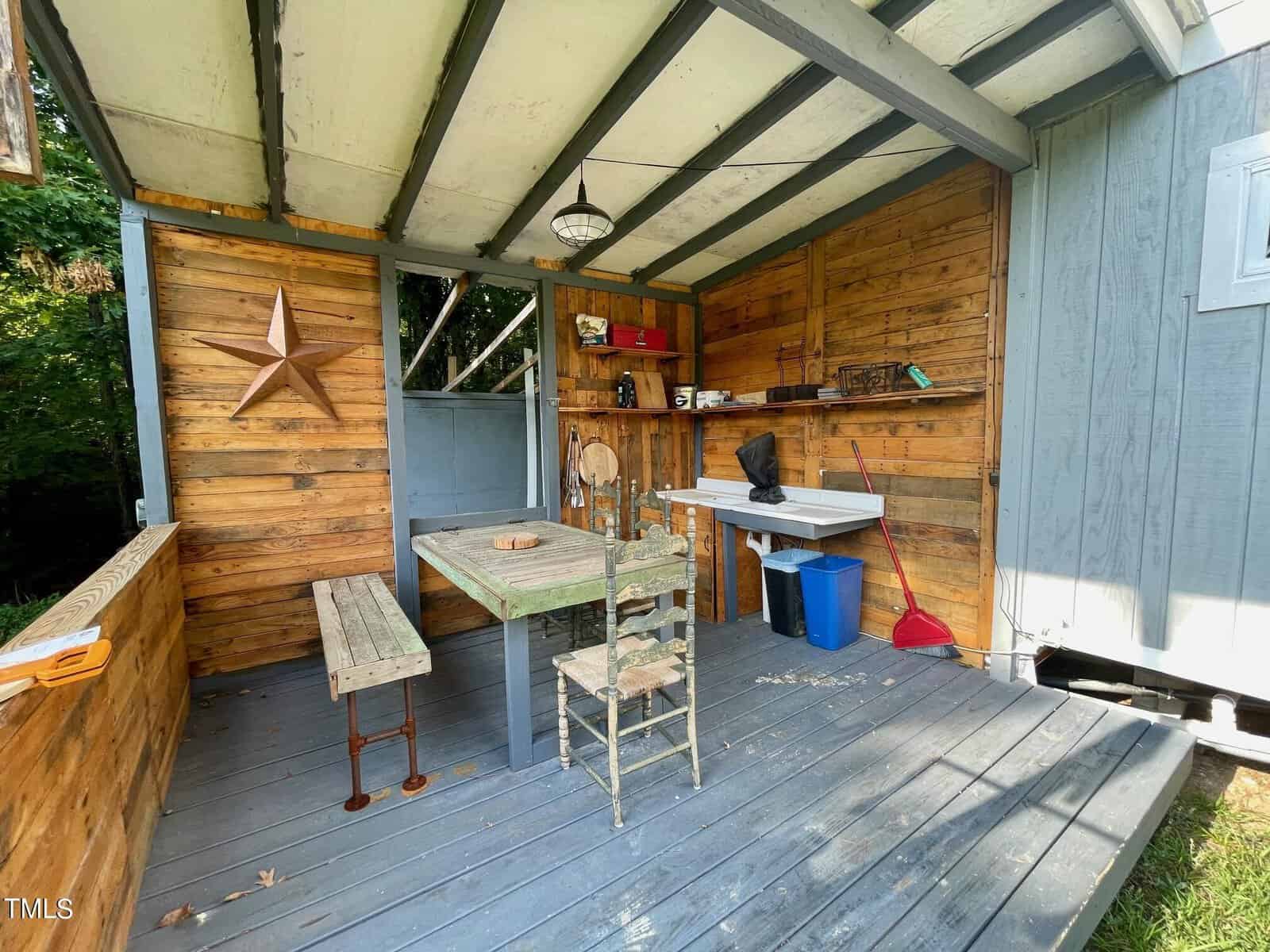 A rustic outdoor shelter with a wooden table and two chairs. The wooden walls feature a large star decoration. A bench and cleaning supplies are nearby. The roof is partially covered, and sunlight filters through the trees surrounding the structure.