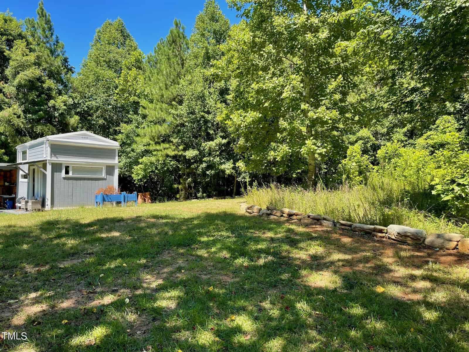 A small, modern shed stands in a sunlit backyard surrounded by tall trees and lush greenery. A patch of wild grass is neatly bordered by rocks, and the bright blue sky is visible above.