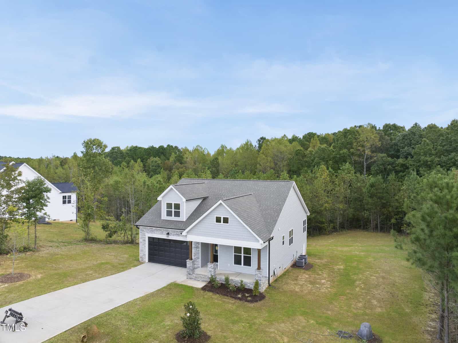 A modern white house with a gray roof, front porch, and double garage sits on a spacious lawn bordered by trees under a blue sky. A concrete driveway leads to the garage.