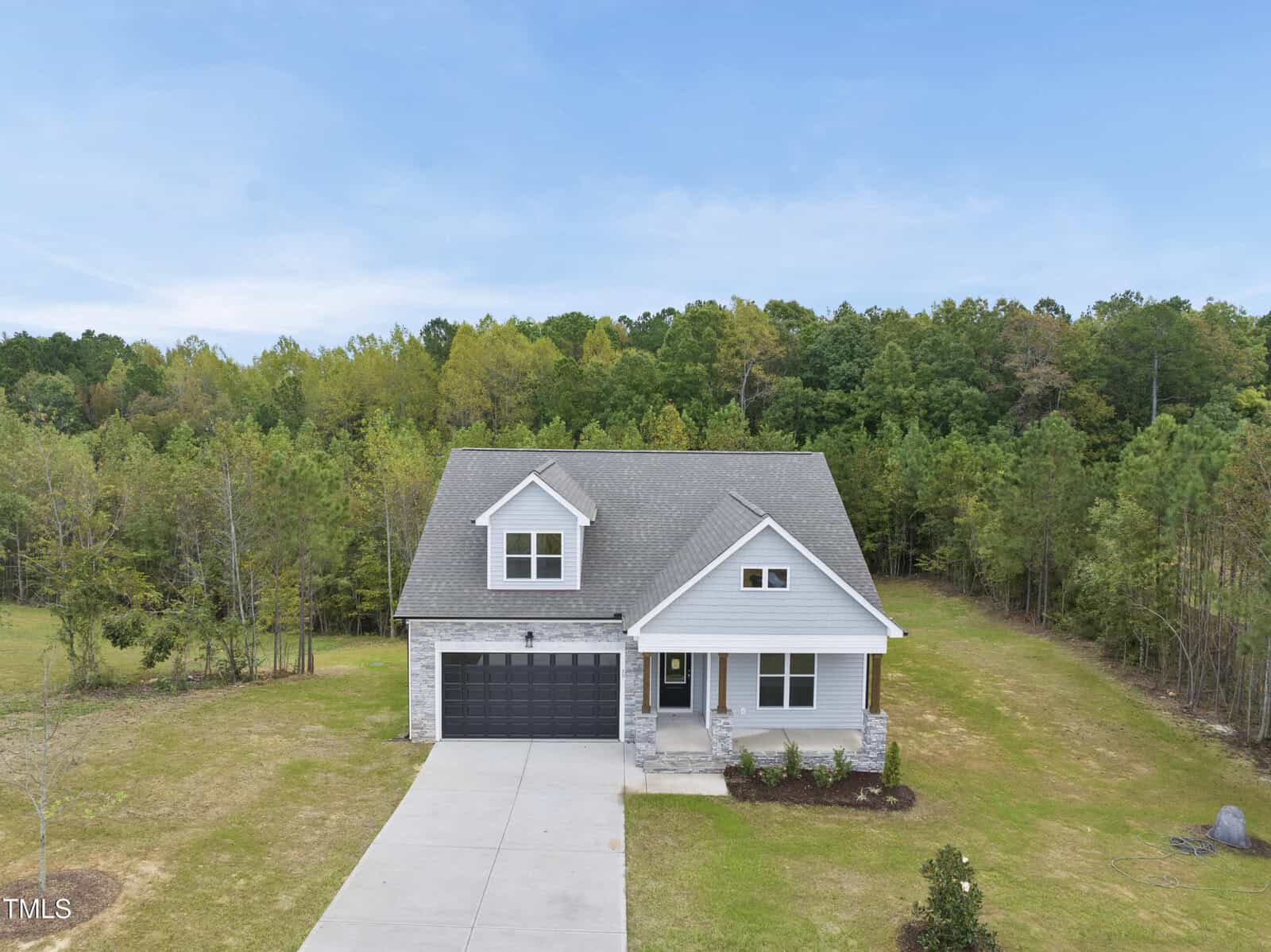 A two-story house with gray siding, a covered front porch, and a black double garage. The home sits on a grassy lot surrounded by trees under a blue sky.