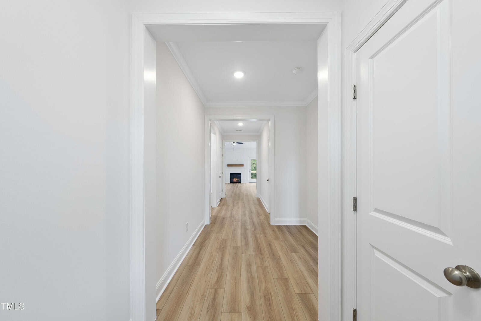 A bright hallway with white walls and wood-style flooring leads to a living area with a fireplace and large windows in the distance. Doors are visible on both sides of the hallway.