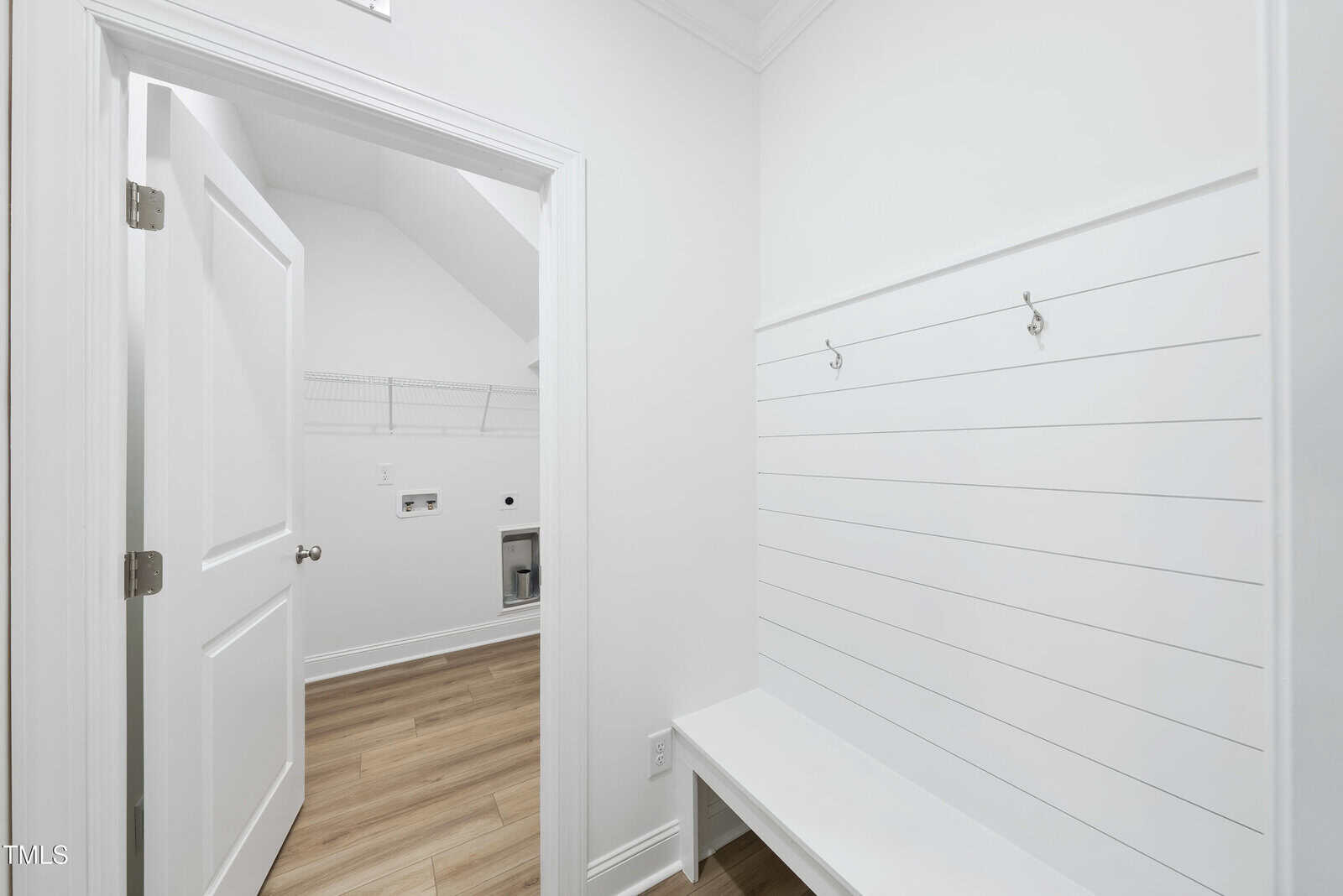 A bright, modern mudroom with white walls, a built-in bench, and wall hooks. The open door leads to a laundry room with wood-style flooring and wall-mounted wire shelving.