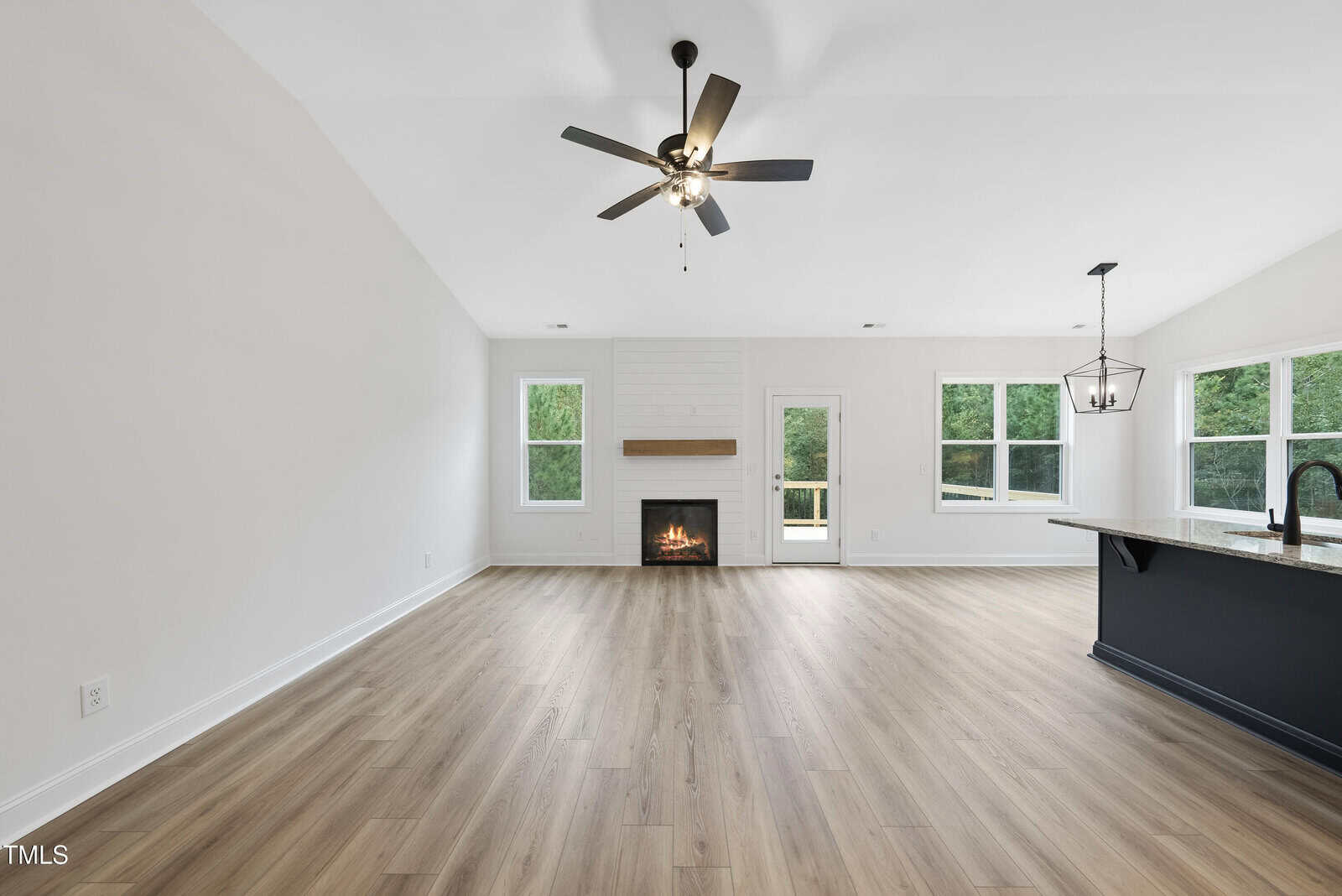 Spacious, modern living area with light wood floors, white walls, a ceiling fan, fireplace, large windows, and a kitchen island with a black faucet and pendant light. French door leads to an outdoor space.