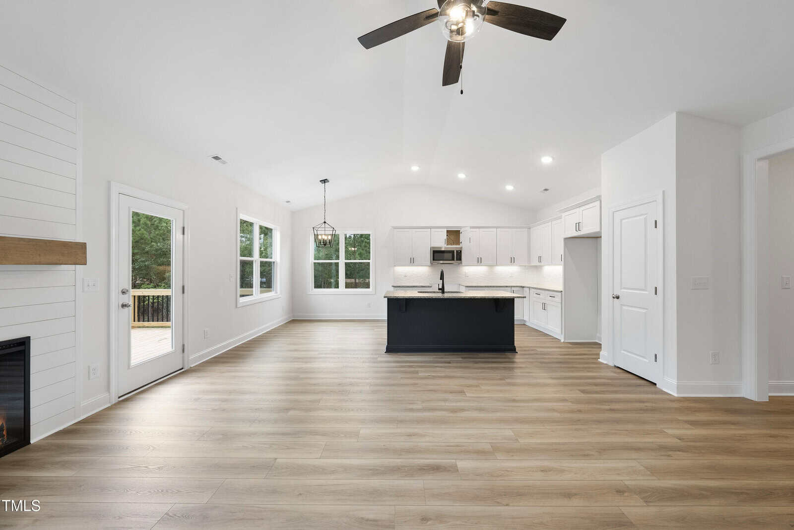 Modern open-concept kitchen and living area with light wood flooring, white cabinets, a black kitchen island, large windows, ceiling fan, and recessed lighting. Glass doors lead to a deck outside.