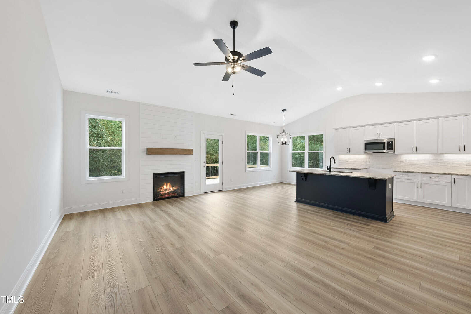 Spacious, modern open-concept living area with light wood floors, a fireplace, ceiling fan, large windows, and a kitchen with white cabinets, island, and stainless steel appliances.