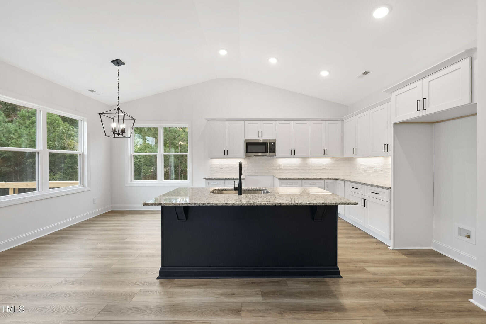 Modern kitchen with white cabinets, light wood flooring, a central black island with a sink, stainless steel appliances, pendant light, and large windows overlooking greenery.