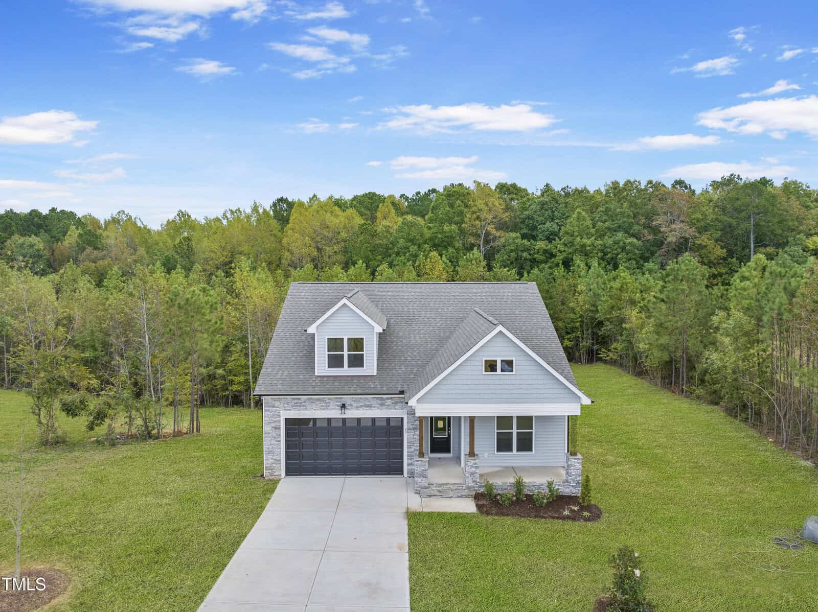 A modern, two-story house with a gray roof and stone accents sits on a large, grassy lot bordered by trees and woods under a blue sky with scattered clouds. A double driveway leads to an attached garage.
