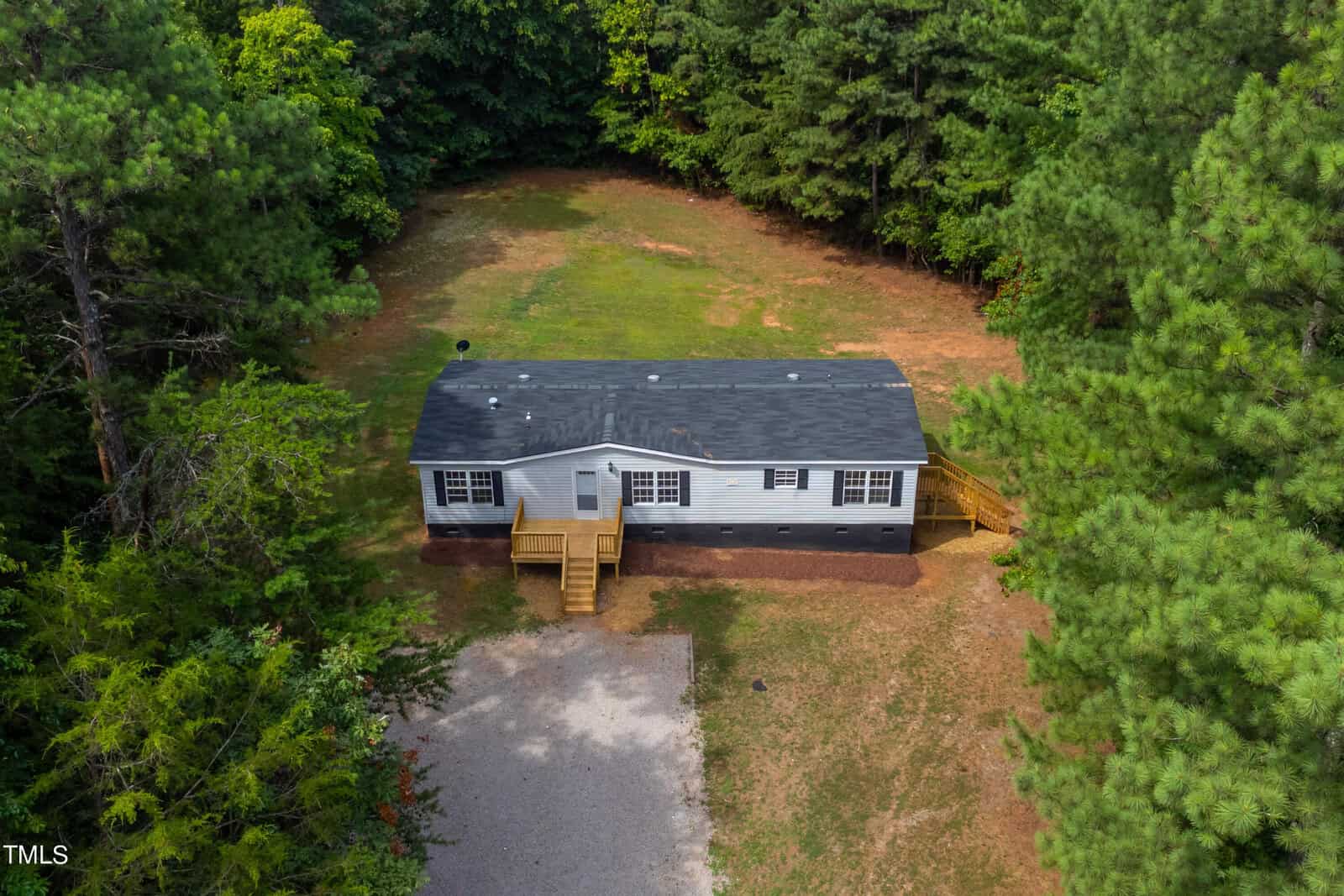 Aerial view of a single-story, light gray mobile home with black roof, situated in a wooded area. The home has a wooden front porch with stairs leading down to a gravel path. The surrounding land is primarily grass, bordered by dense trees.
