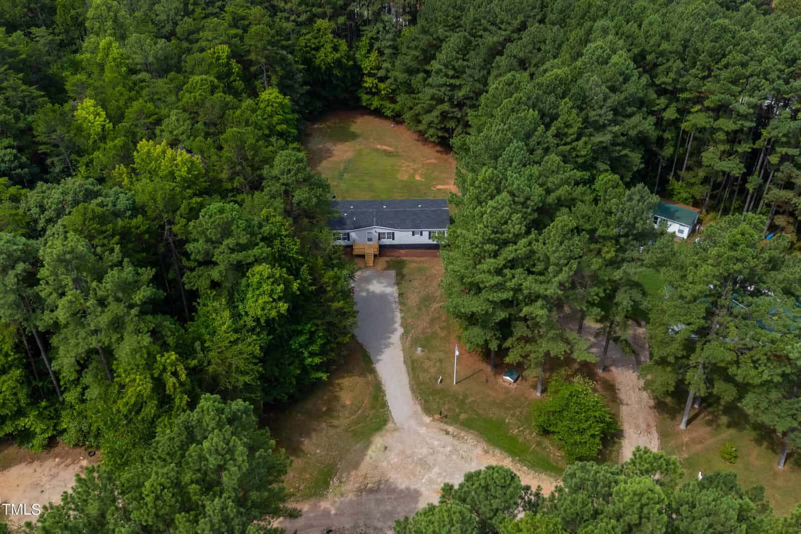 Aerial view of a single-story house with a dark roof surrounded by a dense forest of tall green trees. A driveway curves from the bottom center of the image to the house’s front entrance. A partially cleared yard is visible behind the house.