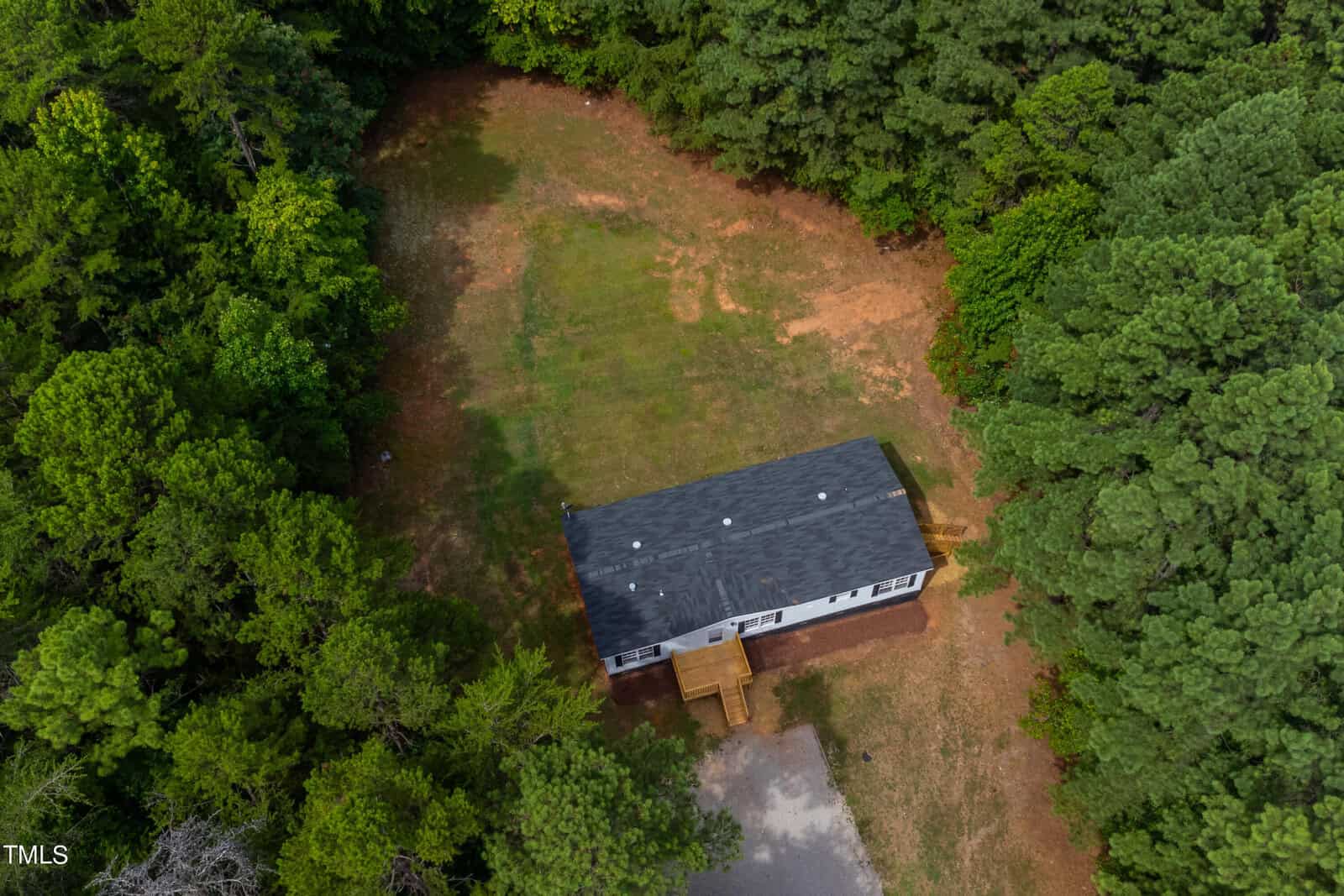 Aerial view of a small white house with a black roof, set in a clearing surrounded by thick green trees. The house has a porch, and the yard shows patches of grass and dirt. The clearing is bordered by dense forested areas.