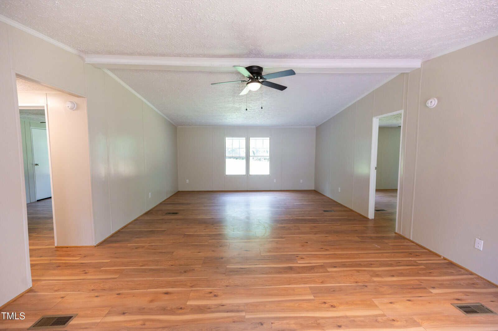 A spacious, empty living room with light wooden floors and beige walls. There are two doorways on the left and right sides leading to other rooms. A ceiling fan with lights is centered on the white, textured ceiling. Two large windows allow ample natural light in.