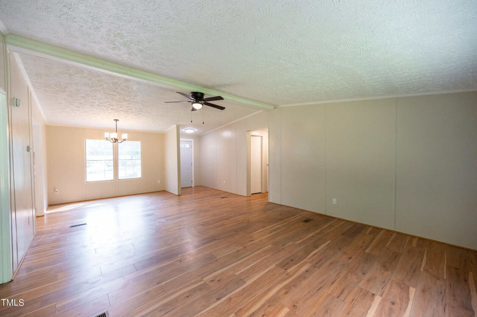 A spacious, empty living room with light-colored hardwood flooring and cream-colored walls. A ceiling fan with lights hangs in the center of the room. There is a large window on the left wall allowing natural light to illuminate the space.