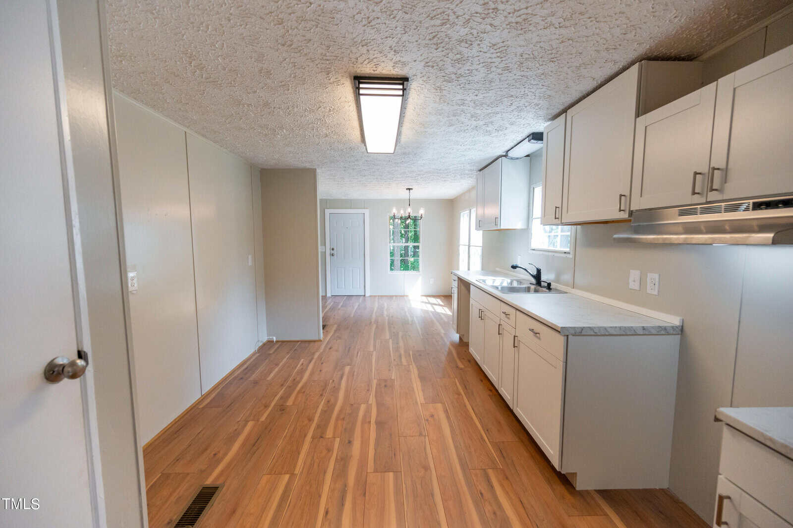 A modern kitchen and dining area with light wood laminate flooring. The kitchen features white cabinets, a white countertop, and a stainless steel sink. A dining space with a hanging light fixture and a door to the outside is visible in the background.