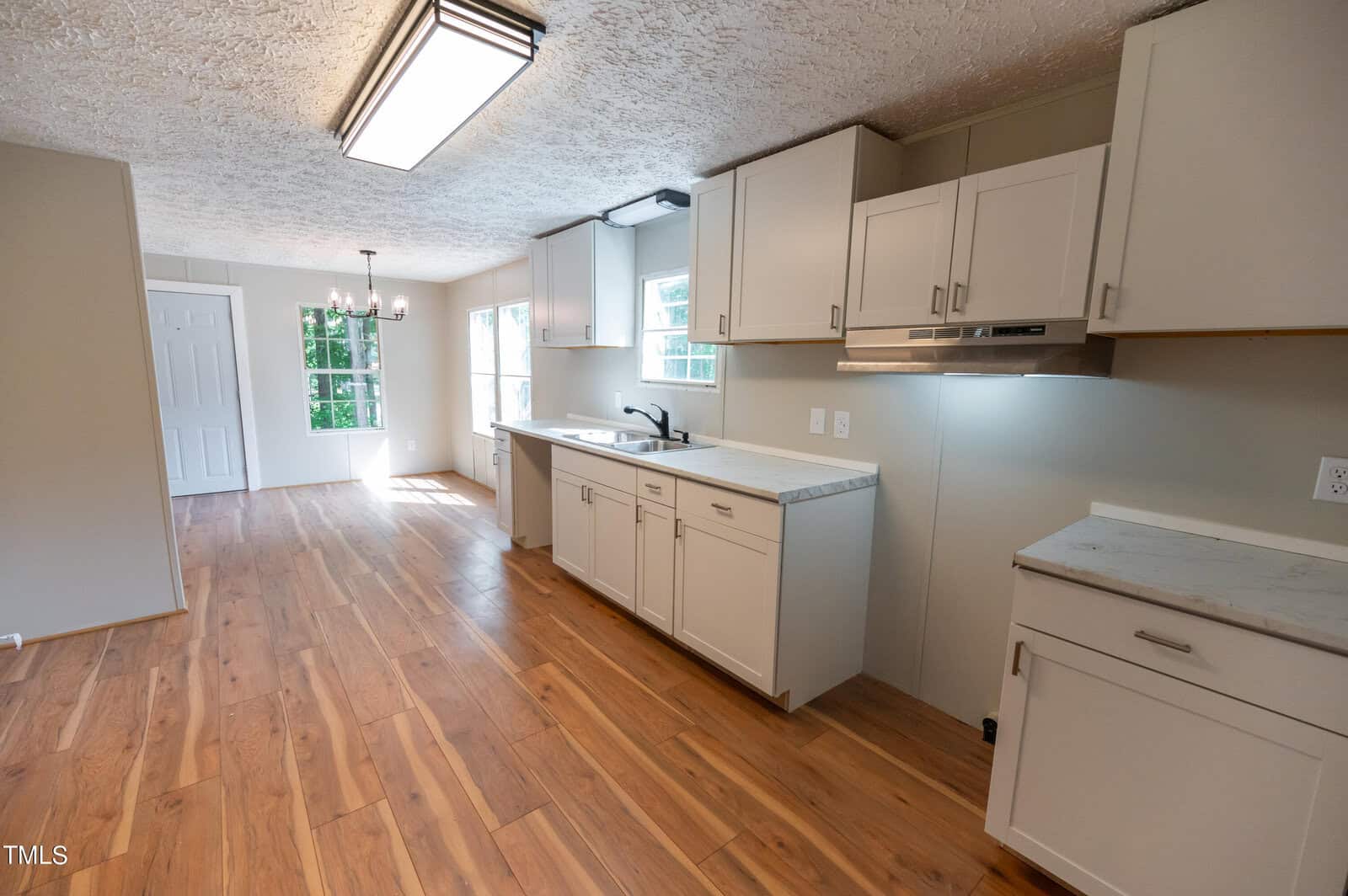 A spacious kitchen with wooden floors, white cabinets, and a sleek black faucet. A window and a glass door on the right let in natural light. The far end of the room features a dining area with a small chandelier, next to a white door.